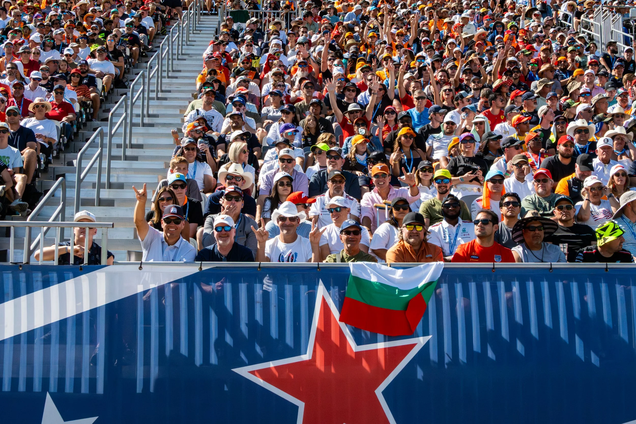 Crowd of people seated in stadium grandstands, some wearing sunglasses and hats, attending an outdoor event, with an Italian flag visible in front.