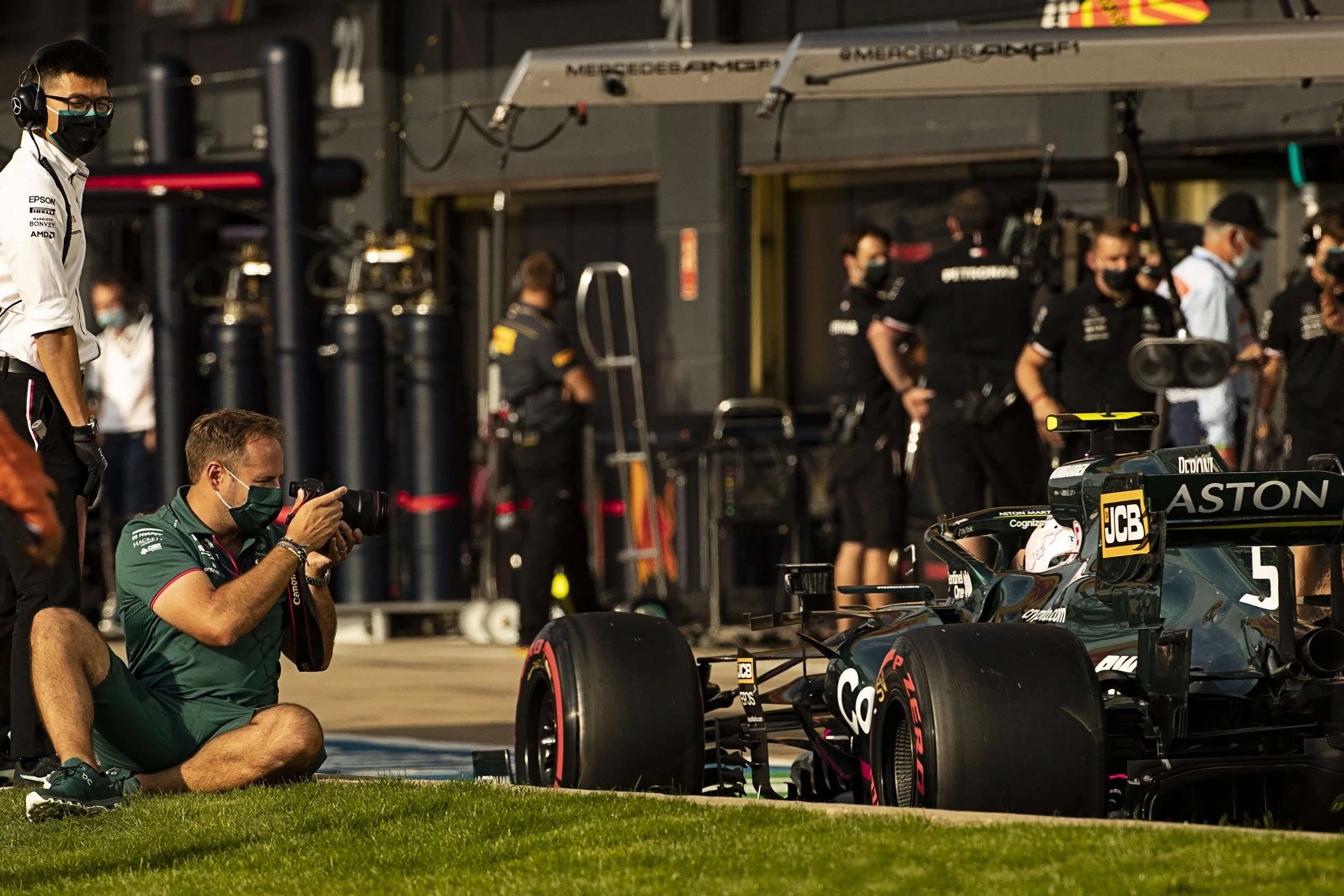 Two Formula 1 race cars in the paddock area, one with Aston Martin branding, surrounded by team members and photographers, with the garage in the background.