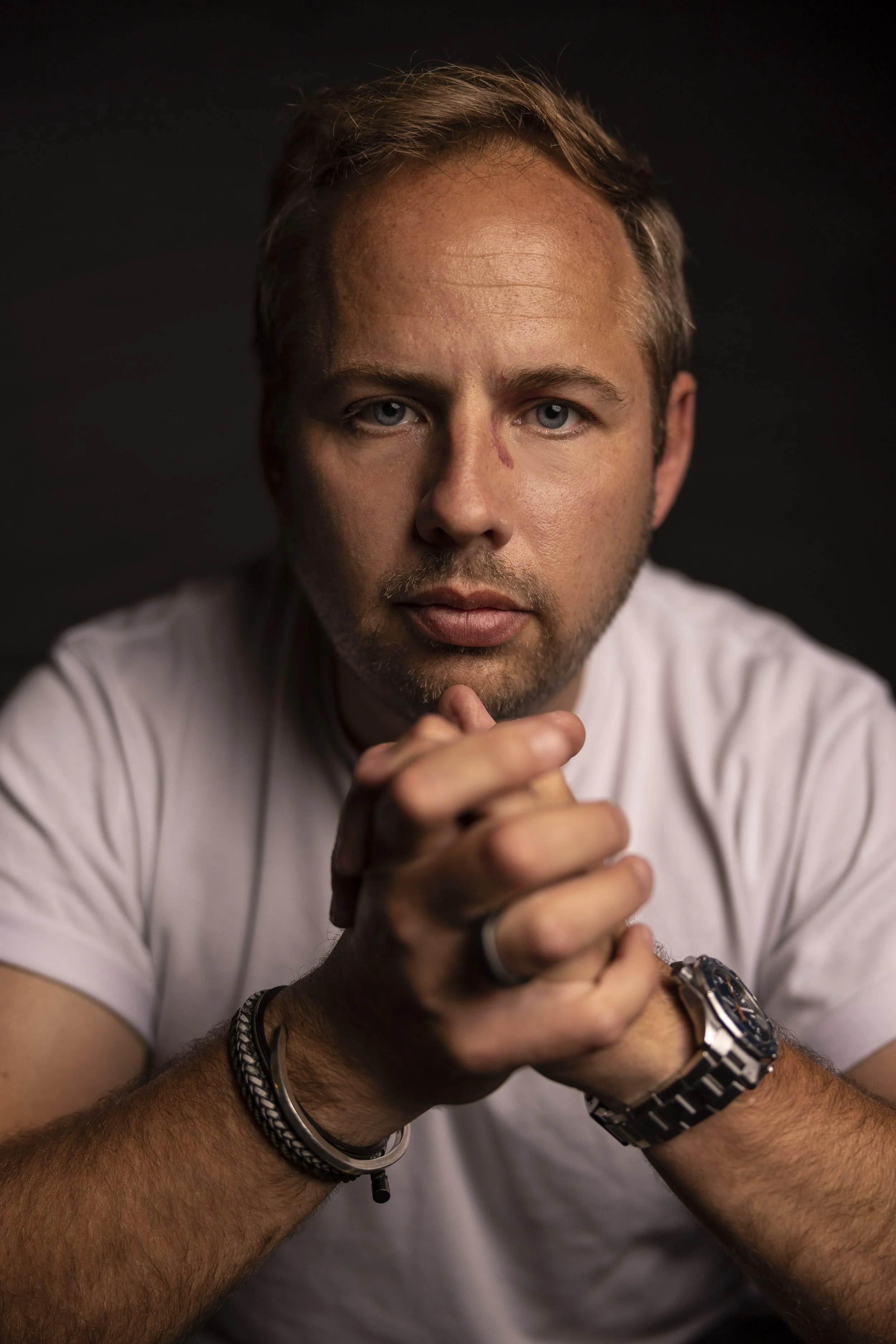 Close-up portrait of a man with blue eyes, short brown hair, and a light beard, wearing a white t-shirt, with his hands clasped and wearing multiple bracelets and a watch, against a dark background.