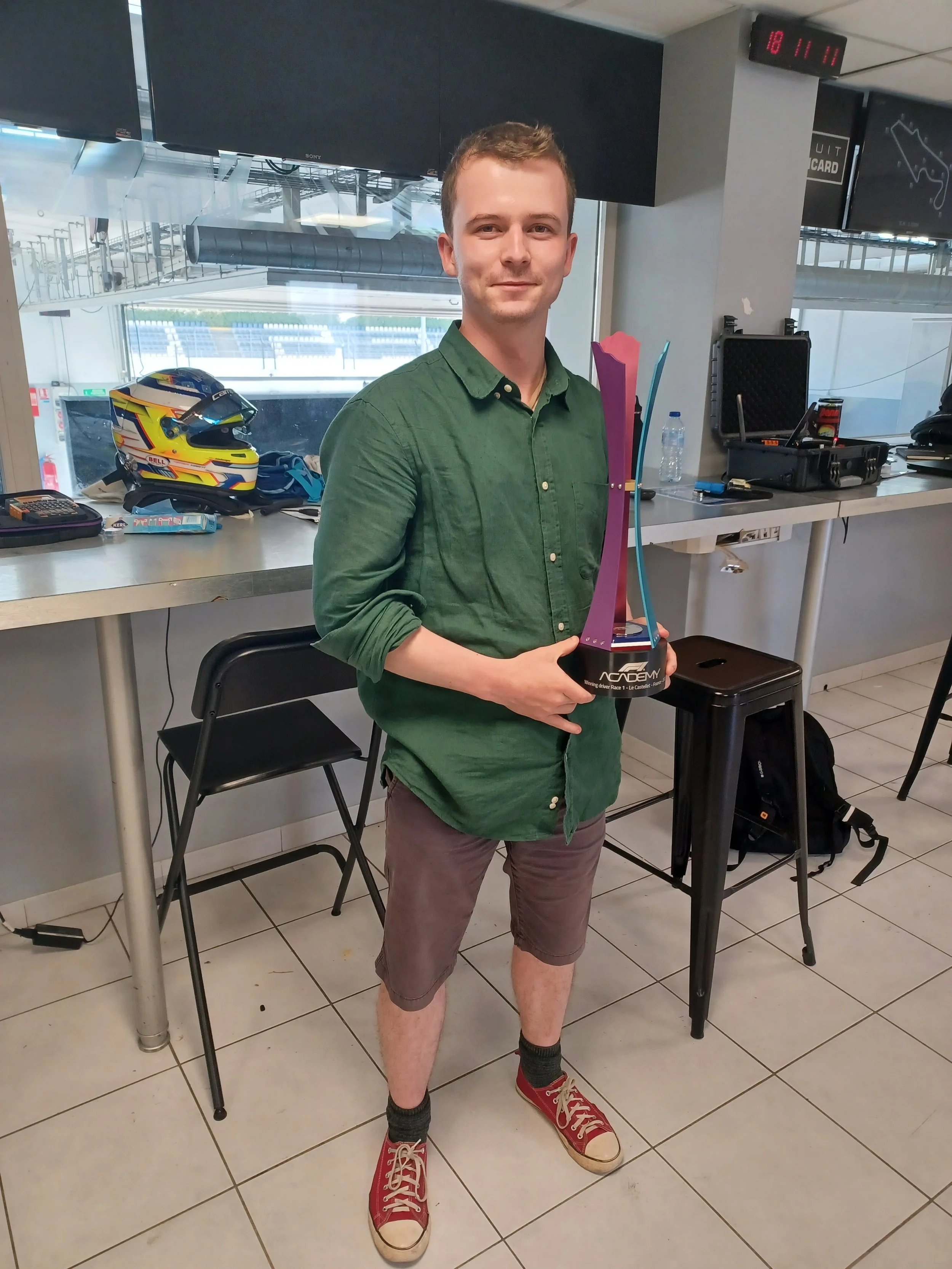 A young man in a green shirt and shorts standing inside a racing venue, holding a racing trophy, wearing red sneakers, with racing helmet and gear on the table behind him.