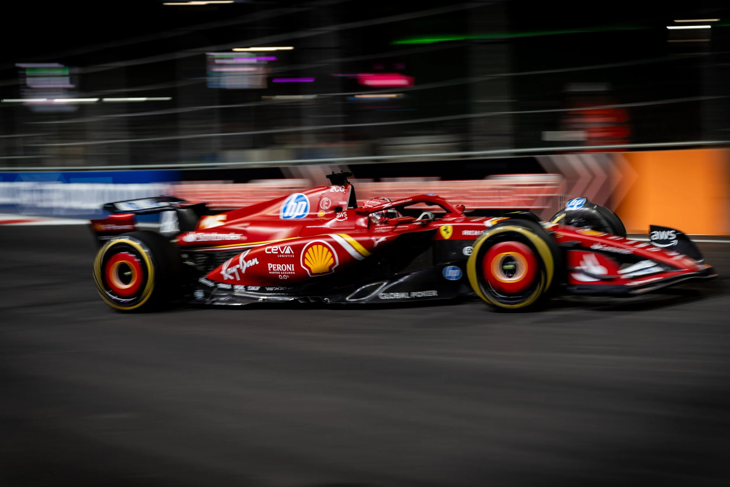 A red Ferrari Formula 1 race car speeds along a race track at night, blurring the background with motion. The car features logos of sponsors like Shell, Ferrari, HP, and AWS.