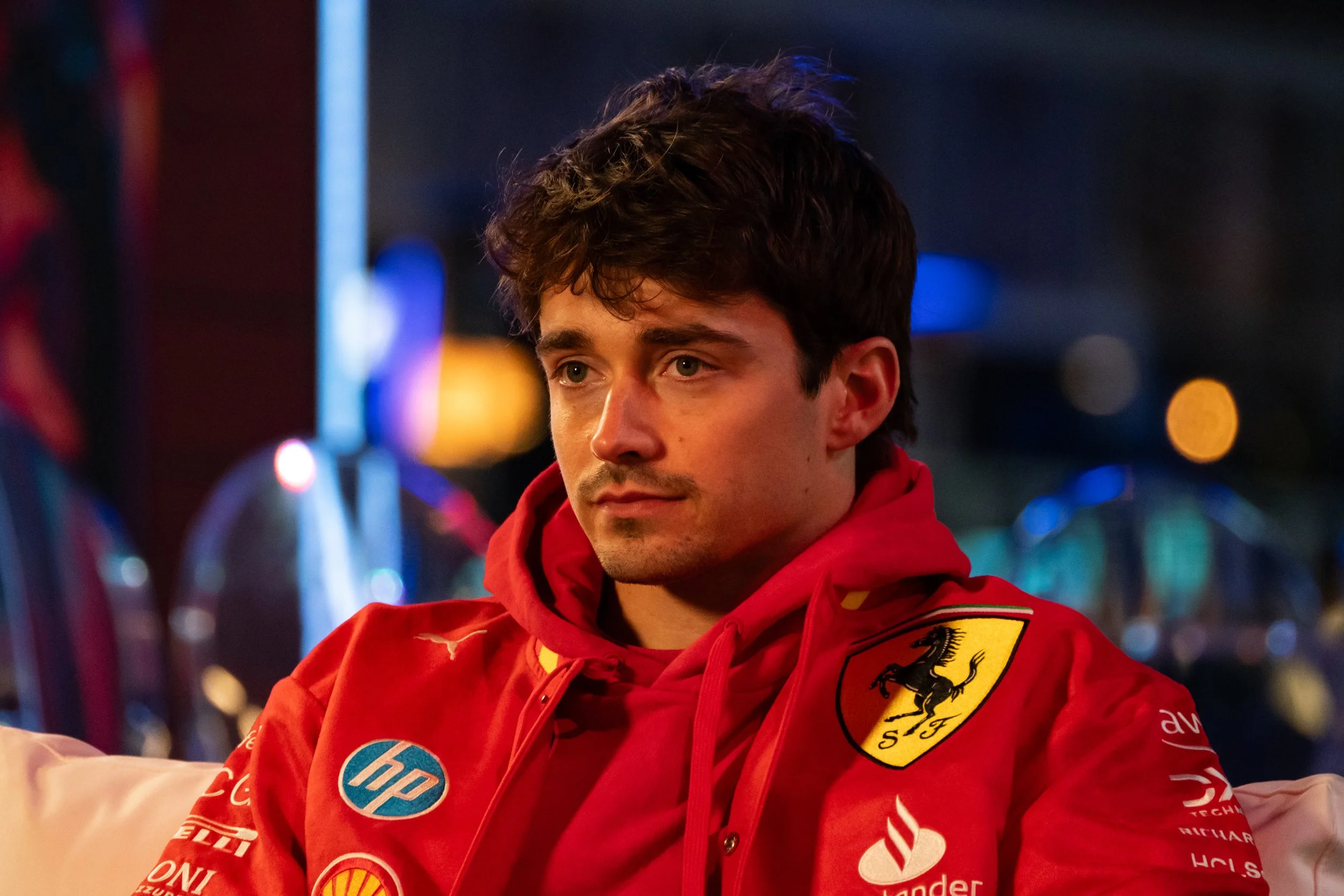 Charles Leclerc wearing a red Ferrari racing jacket with sponsor logos, during the interview at Las Vegas Grand Prix paddock with a beautiful Las Vegas night vibes onb a background.