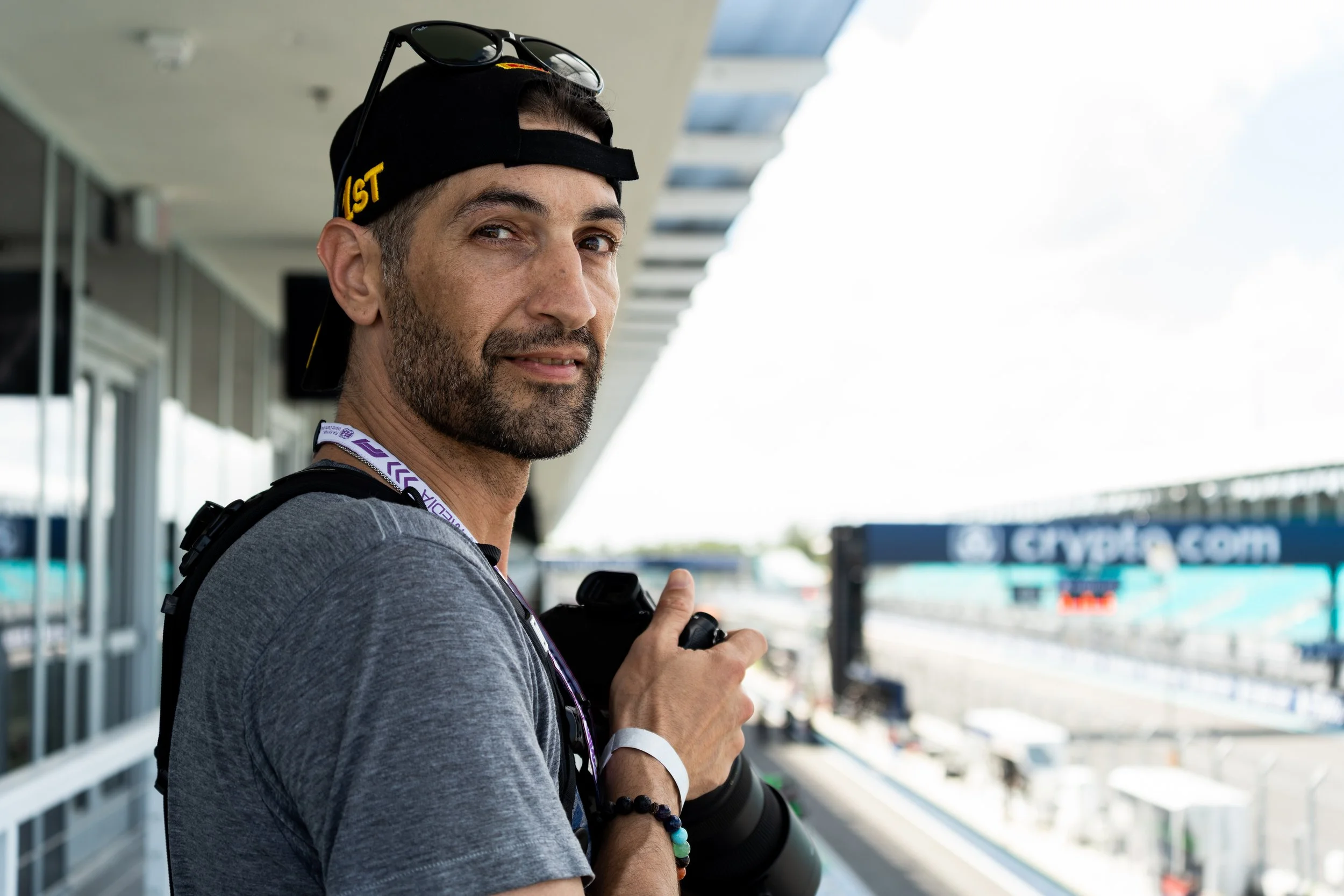A man in a gray shirt and black cap with sunglasses on top, holding a camera at a racing event, standing in front of a grandstand with a partly cloudy sky.