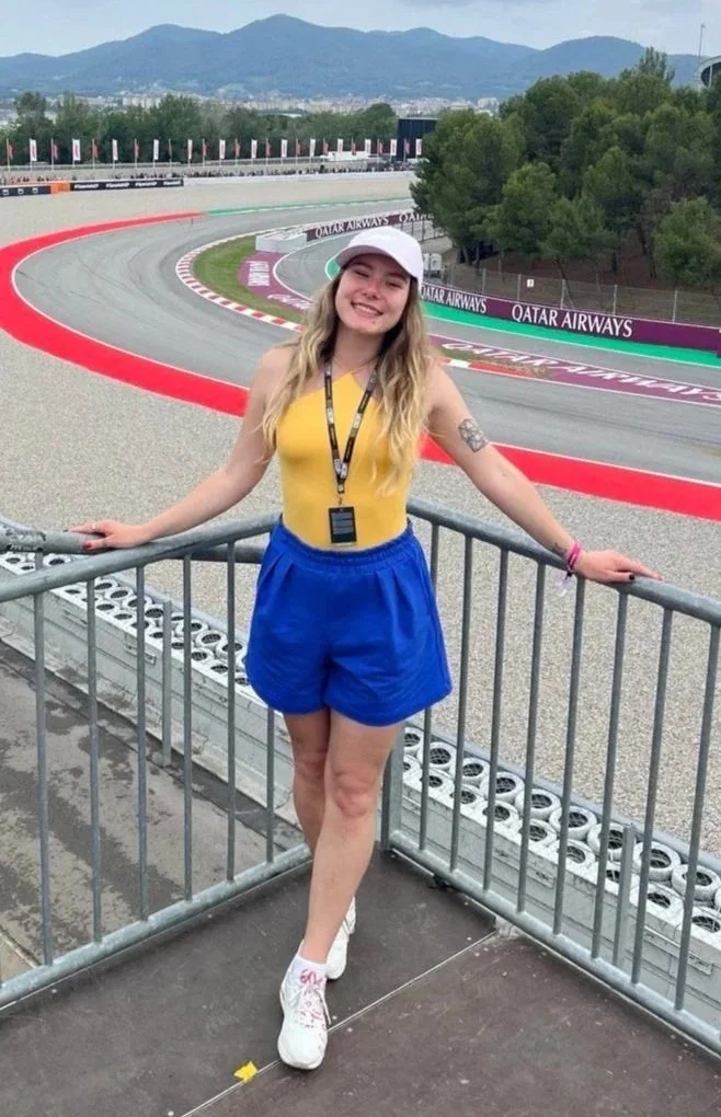 Anna Kharchenko on a viewing platform at a race track with her arms resting on the railing, smiling at the camera. The race track is empty, with red and white curbing and advertising banners, and mountains and trees in the background.