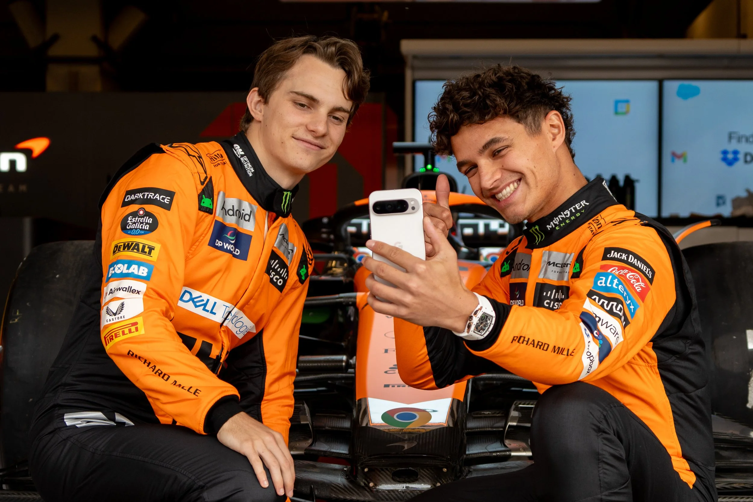 Oscar Piastri and Lando Norris in front of McLaren Formula 1 car at COTA in Austin, Texas, smiling at a smartphone, in a garage or paddock area.