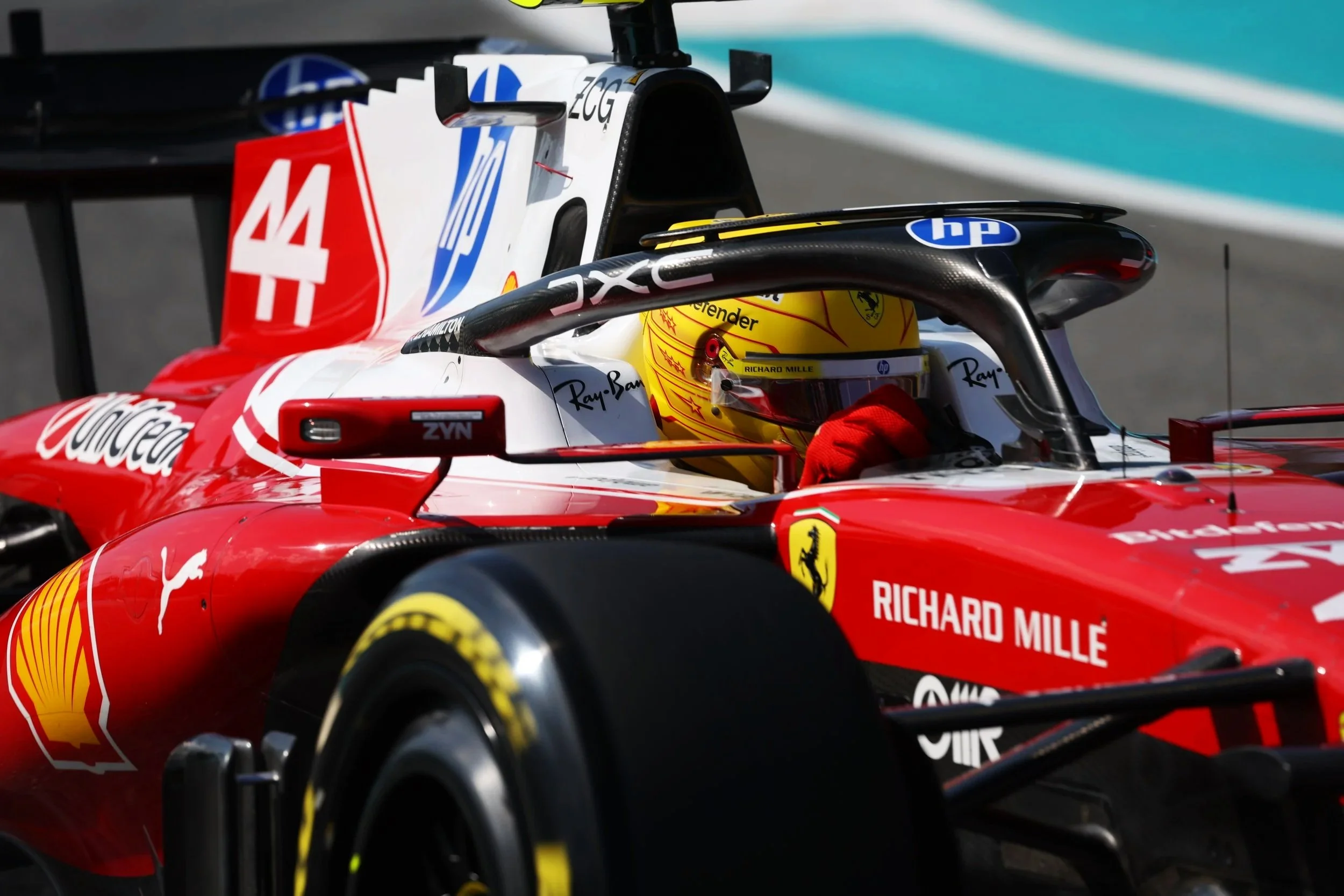 Close-up of a Ferrari Formula 1 car with a driver inside, wearing a yellow helmet. The car features sponsorships from Coca-Cola, Shell, HP, and Richard Mille, with a prominent red and white color scheme and the number 44 on the rear wing.