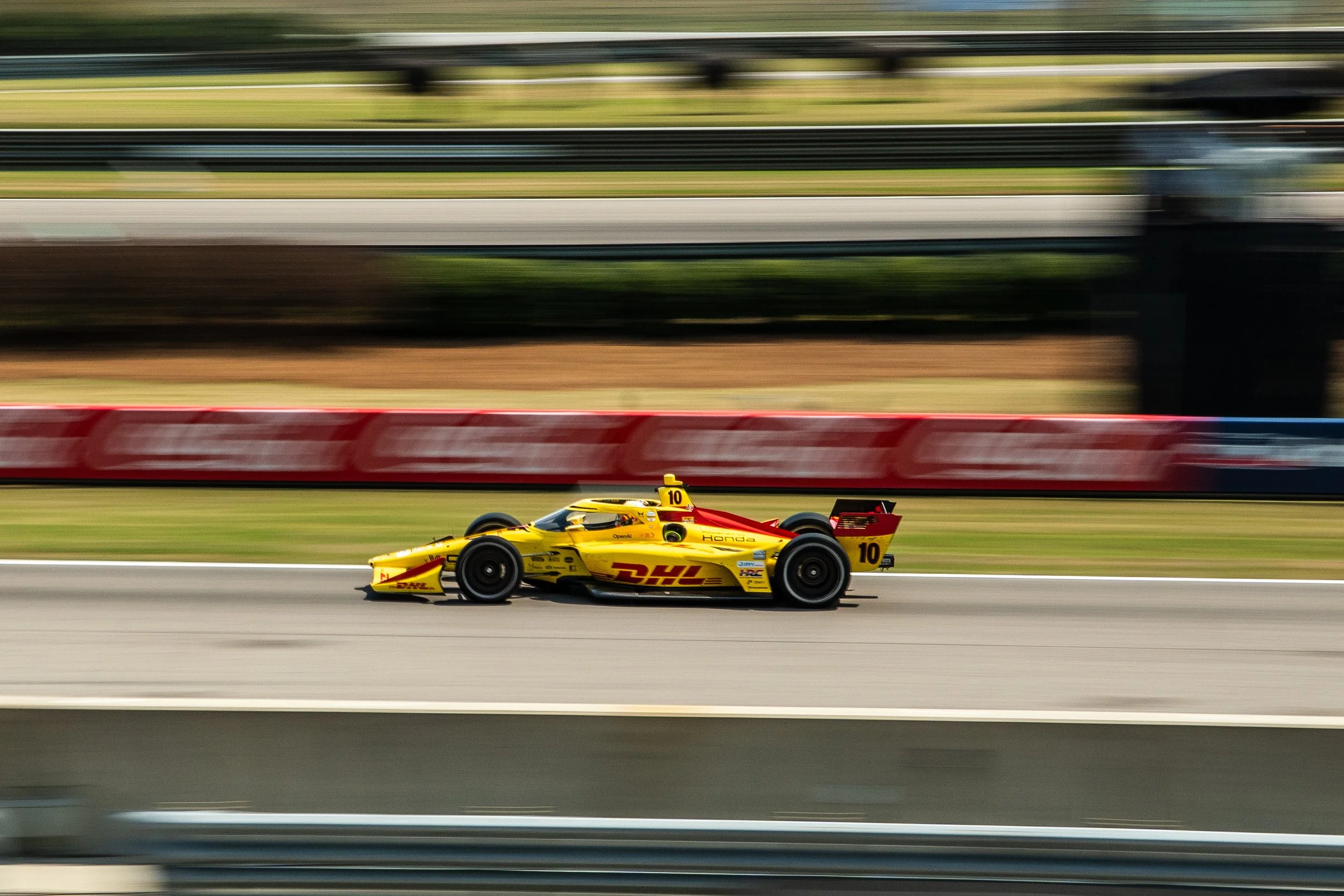 A yellow open-wheel race car with DHL branding on the side, numbered 10, speeding on a racetrack with blurred background.