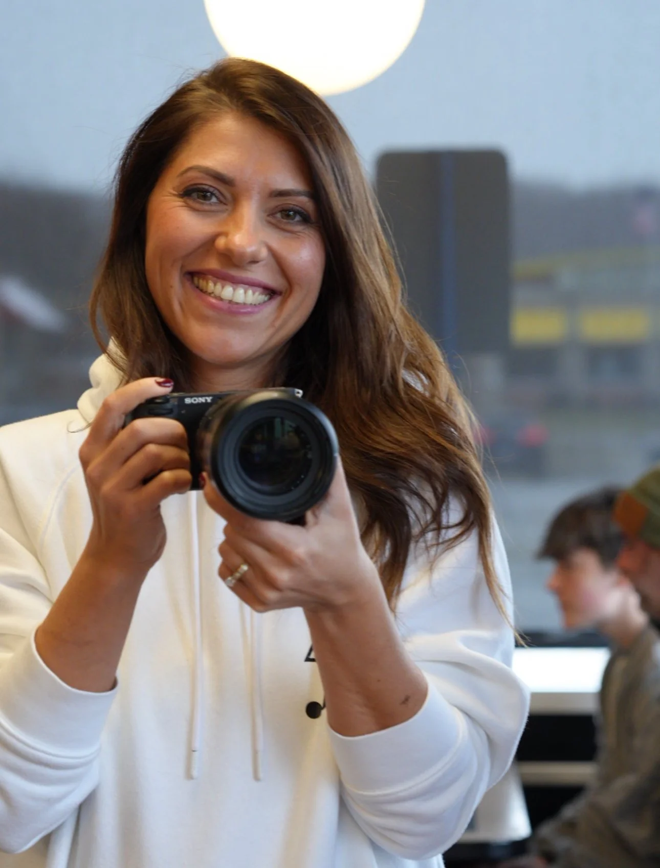 Olga Ganay smiling and holding a camera indoors, with other people working at desks in the background.