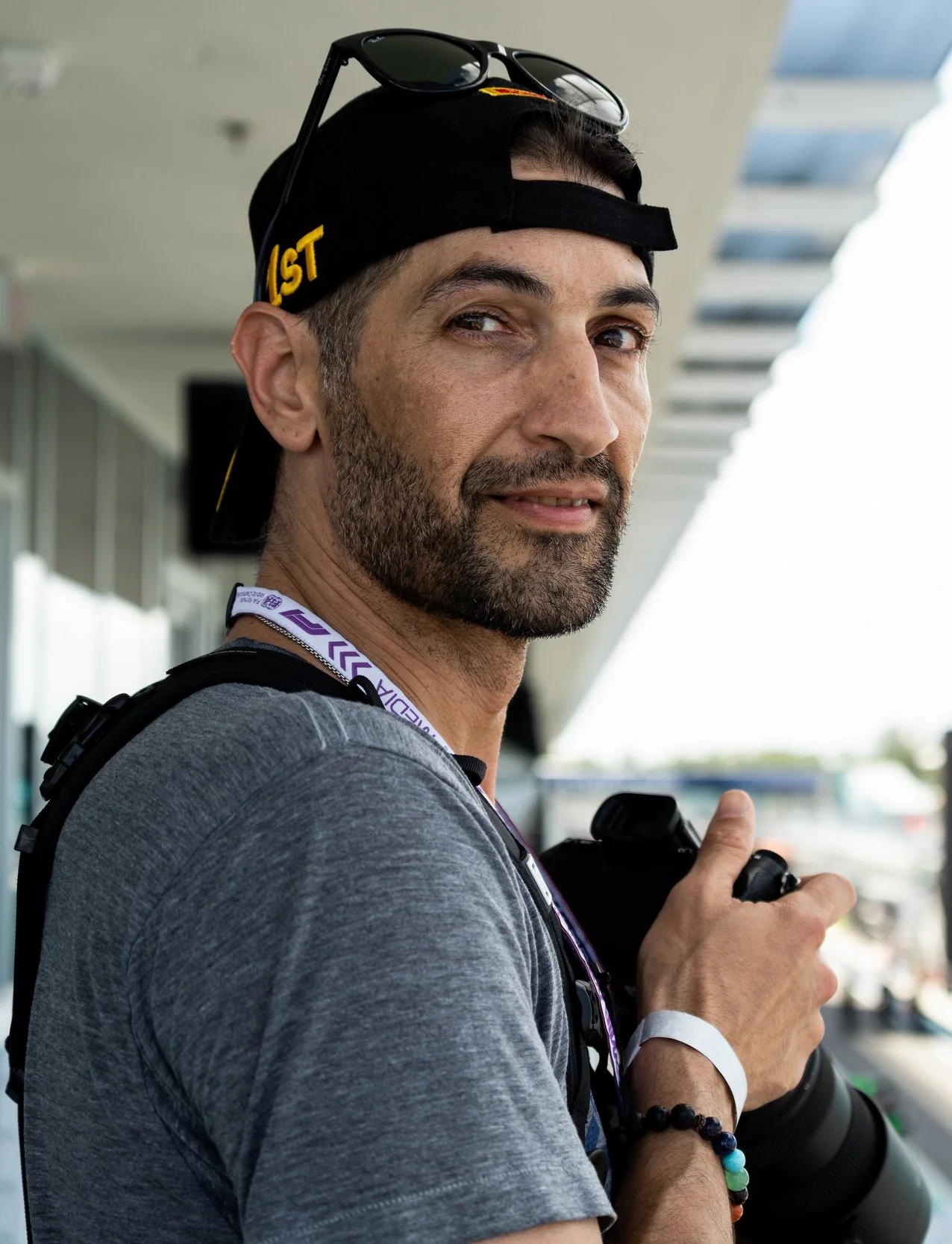 A man with a beard wearing a black cap backwards with sunglasses on top, a gray t-shirt, and a camera strap around his neck, standing outdoors near a modern building with large windows and metal architecture, giving a thumbs up.