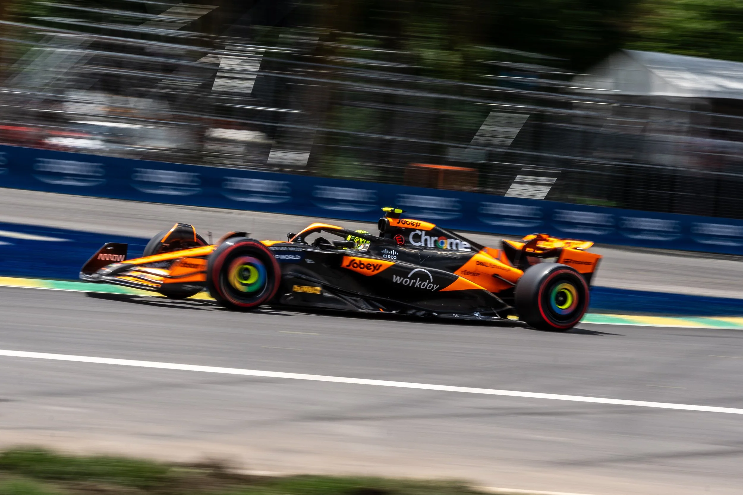 A McLaren papaya race car in orange and black speeds along a racing track, with motion blur in the background during Canadian GP.