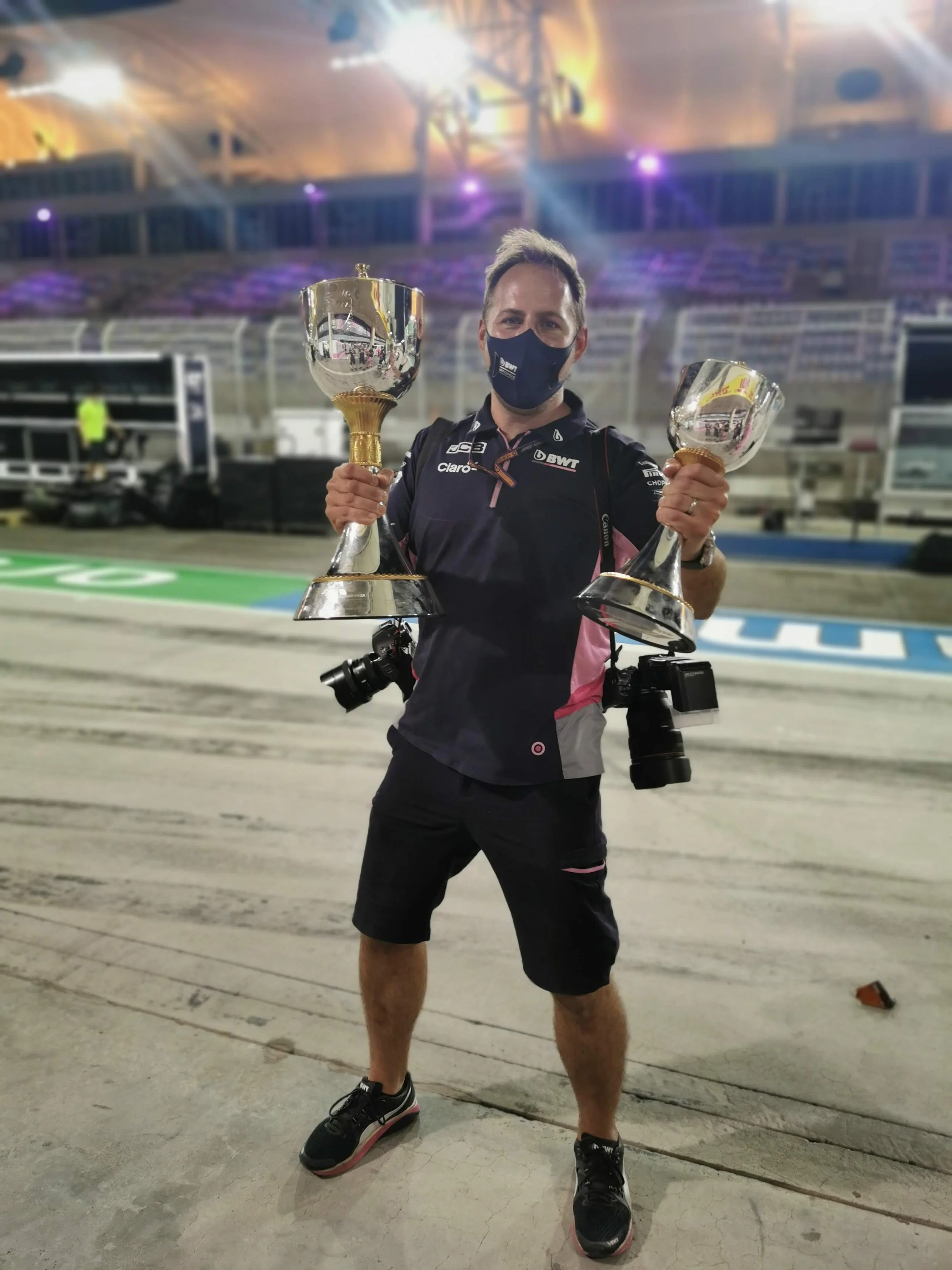 A man wearing a face mask and black athletic shorts stands on a race track holding two silver trophies, with cameras hanging around his neck and photography equipment on his waist in the background.