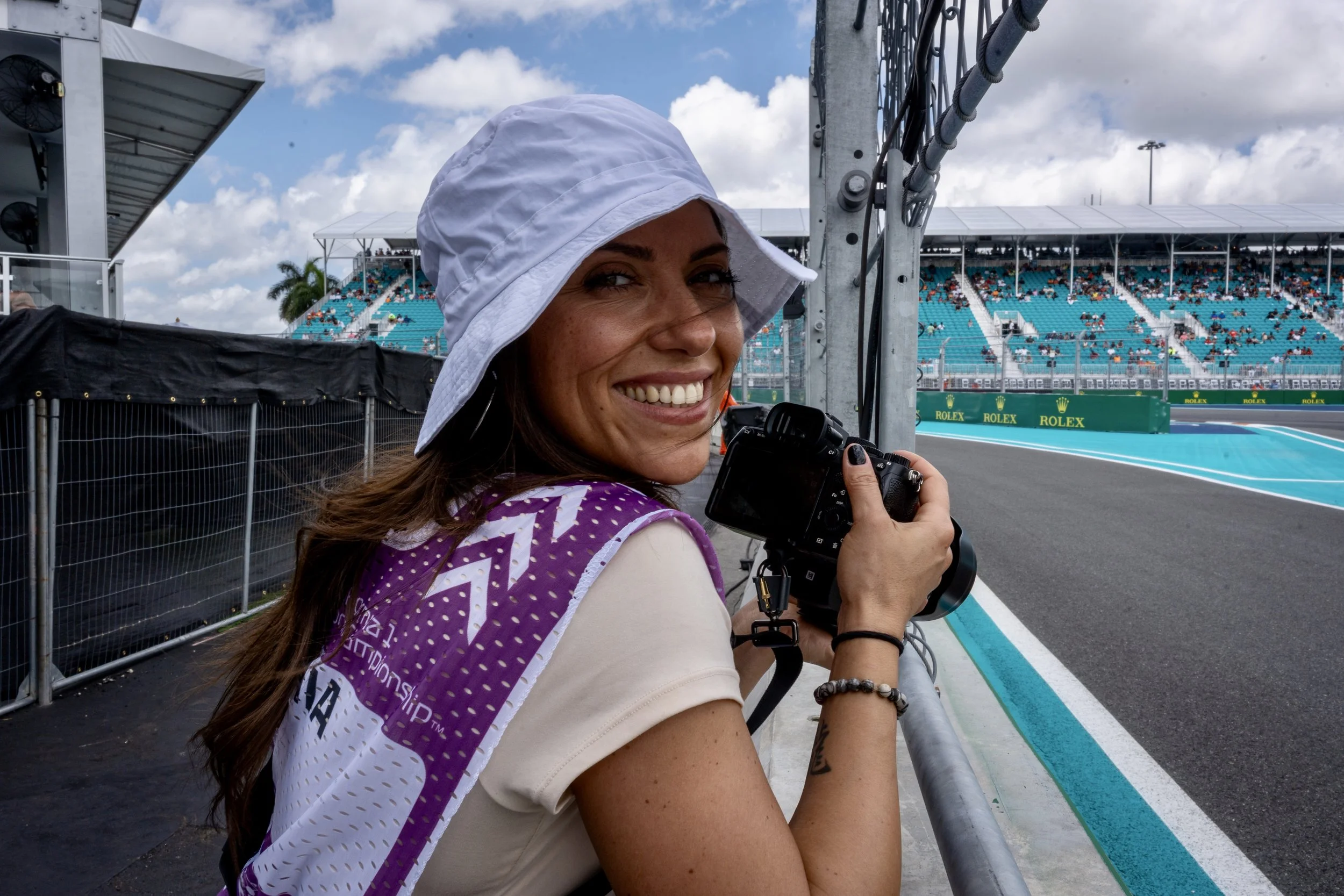 A woman smiling, wearing a white sun hat, holding a camera, sitting at a race track with spectators in the stands in the background under partly cloudy skies.
