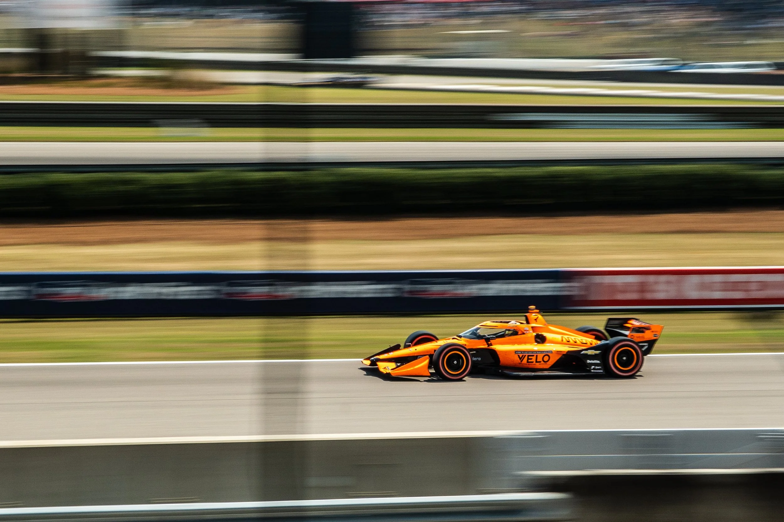 A bright orange race car driving on a race track at high speed, with motion blur in the background.