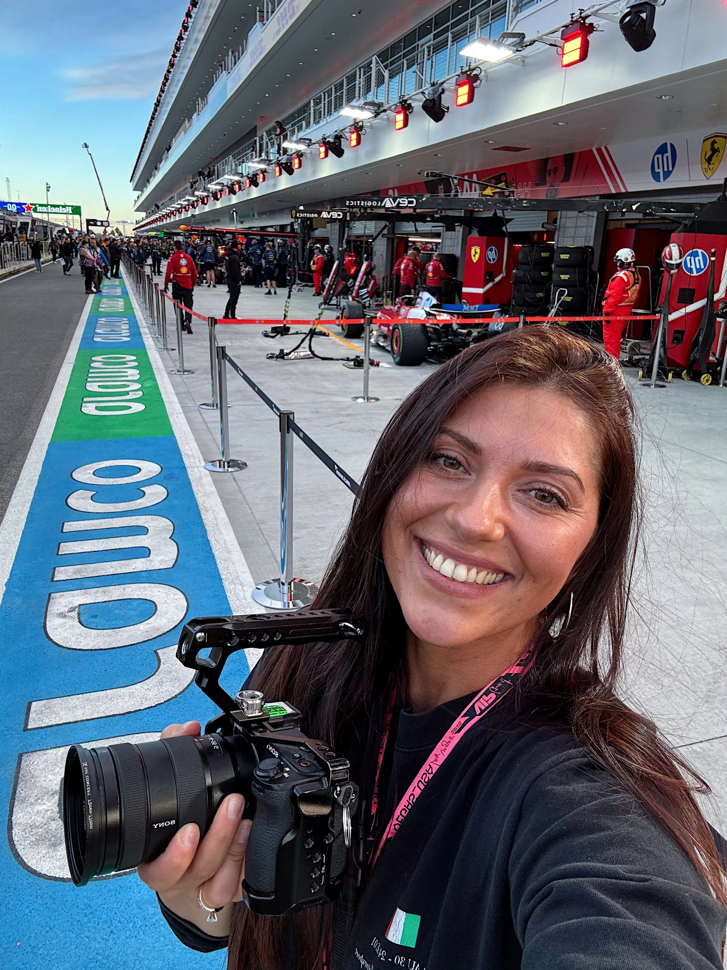 Olga Ganay smiling and holding a camera at a racing track with Formula 1 cars in the garage and people walking around in the background.