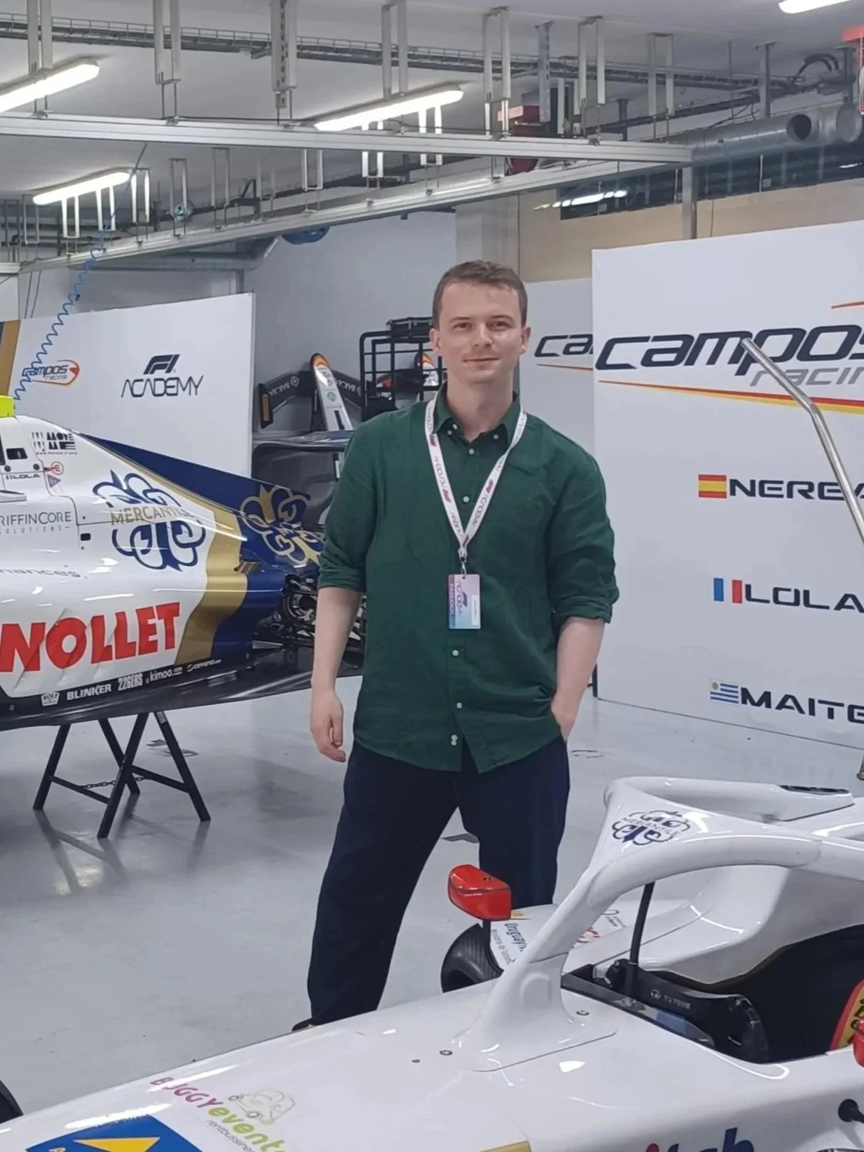 A man in a green shirt standing in a garage with a Formula 1 racing car and banners with racing team logos behind him.