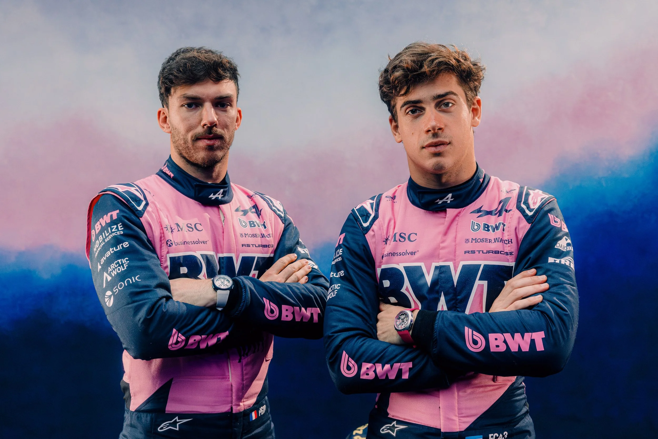 Two BWT Alpine Formula One drivers pose in pink and blue race suits against a pink and blue smoke background.