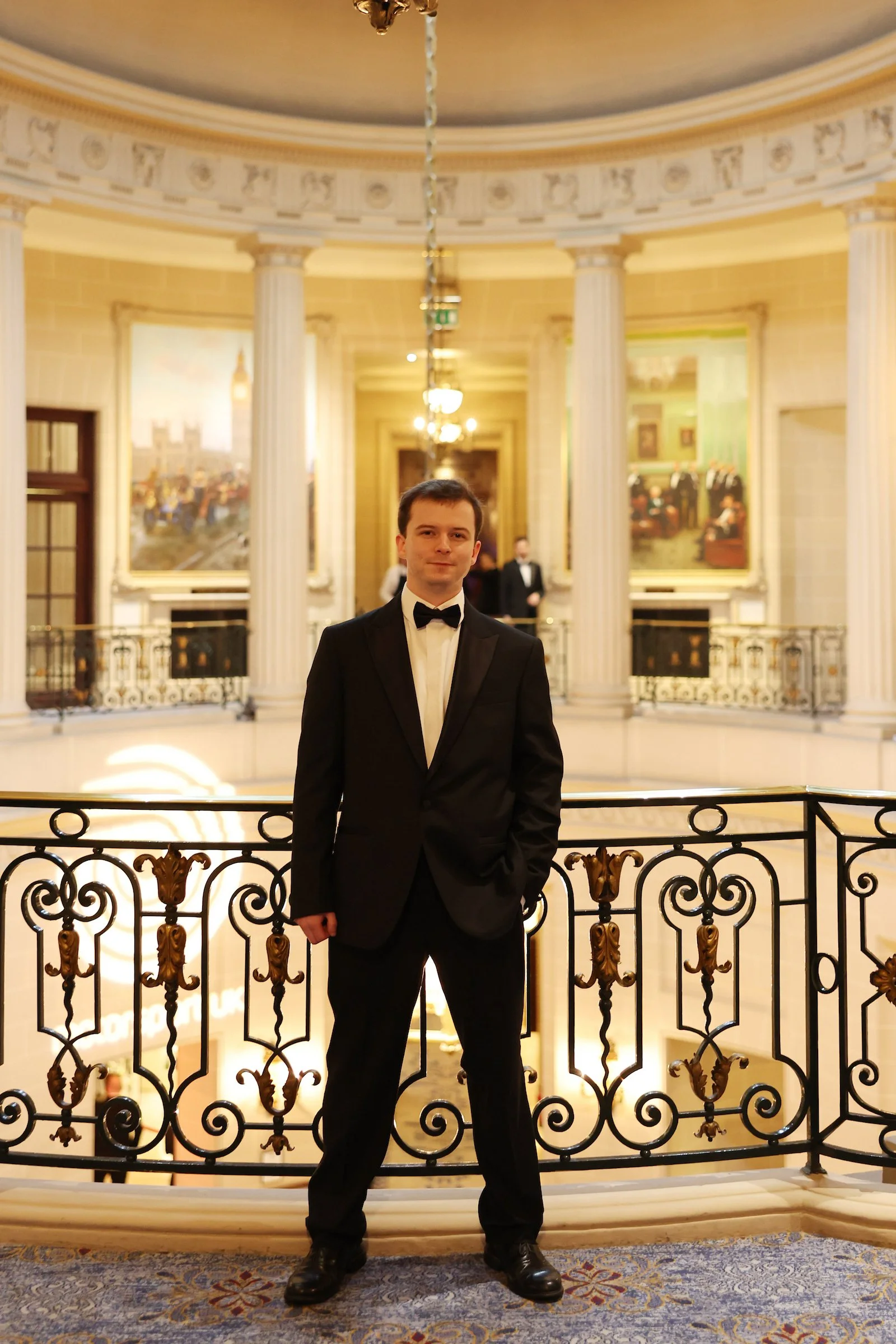 Thiemo Albers-Daly in a black tuxedo with a bow tie, standing in front of an ornate railing in an elegant, historic building with classical architecture. The background features tall columns, large paintings, and warm lighting.
