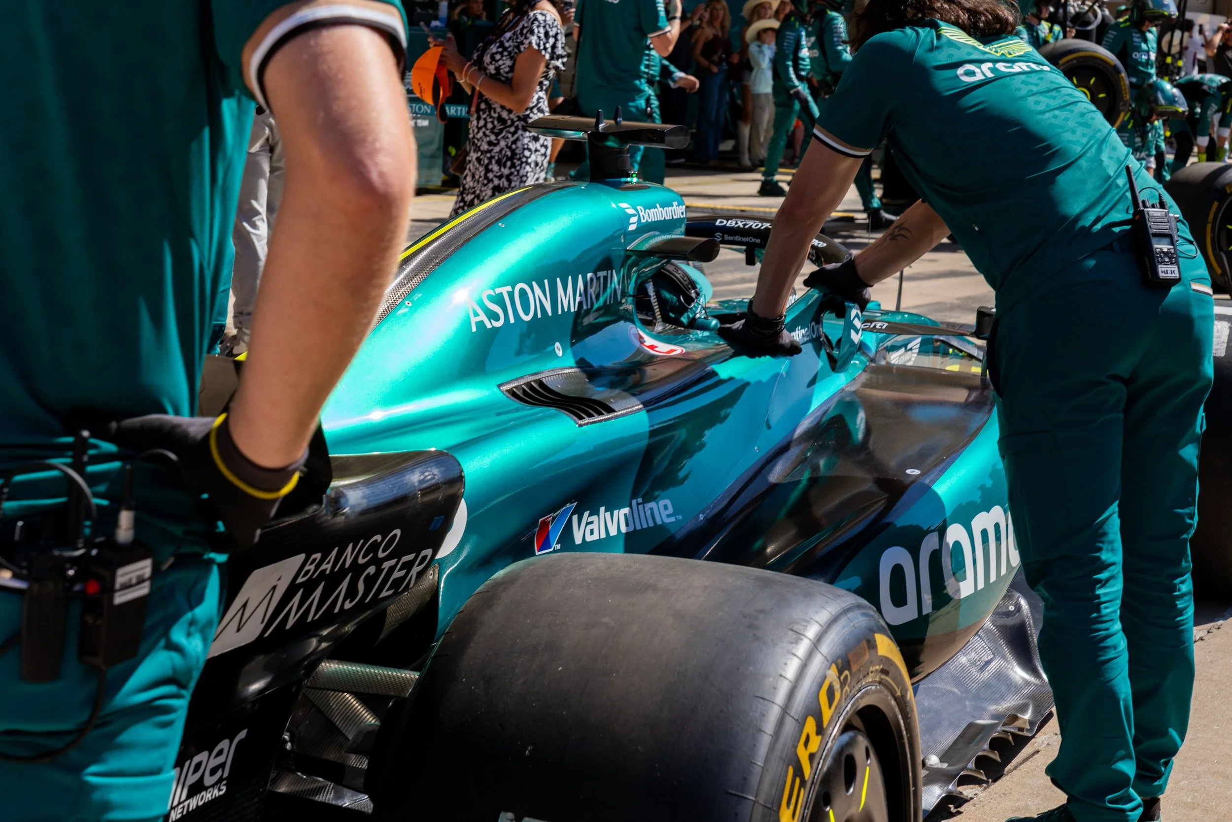 Aston Martin F1 car being serviced by mechanics in the pit lane, with a crowd in the background. The car is teal with sponsorship logos, including Aston Martin and Valvoline.