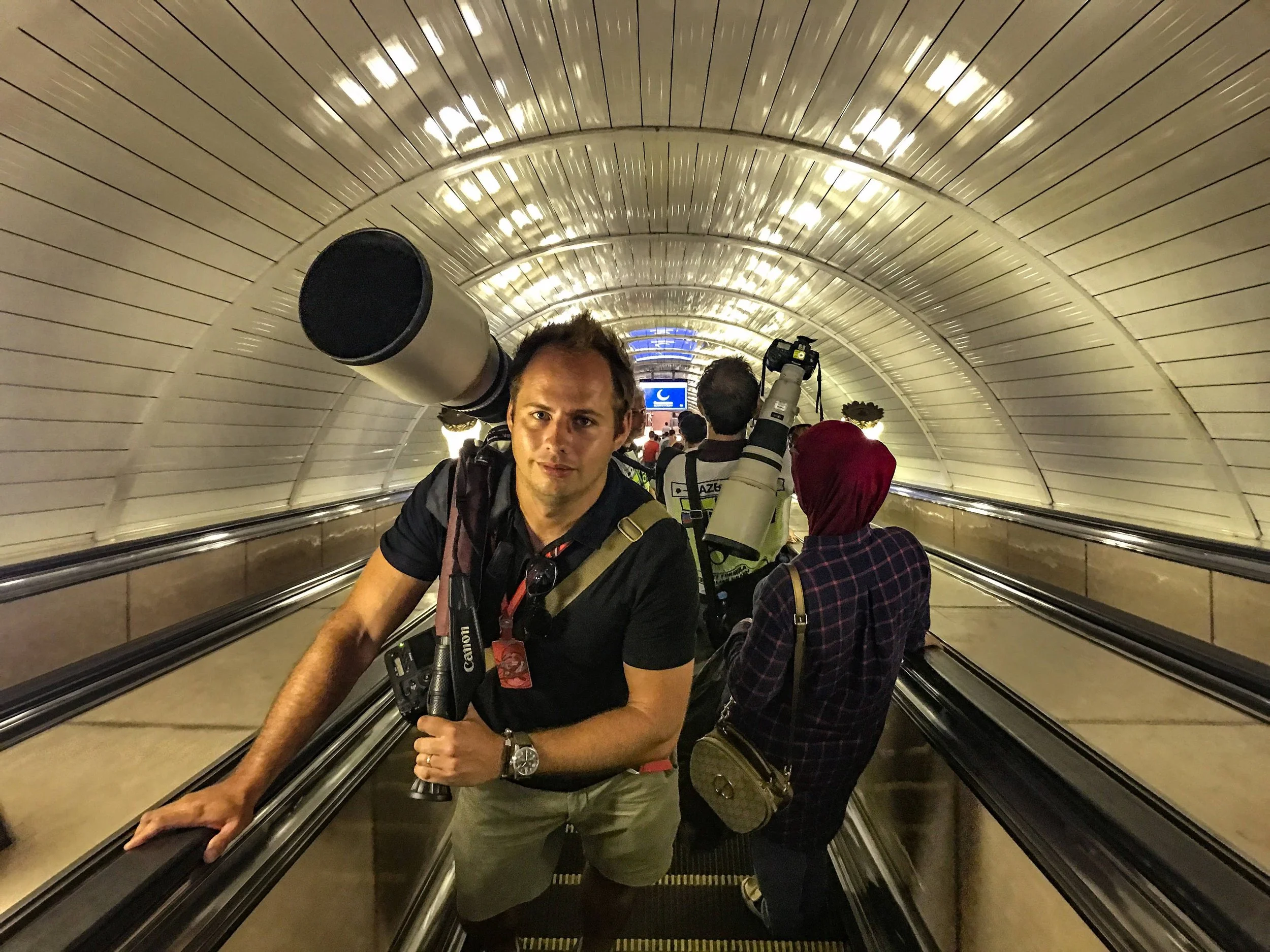 People walking through a curved underground tunnel with metallic walls, carrying various equipment including cameras and telescopes.