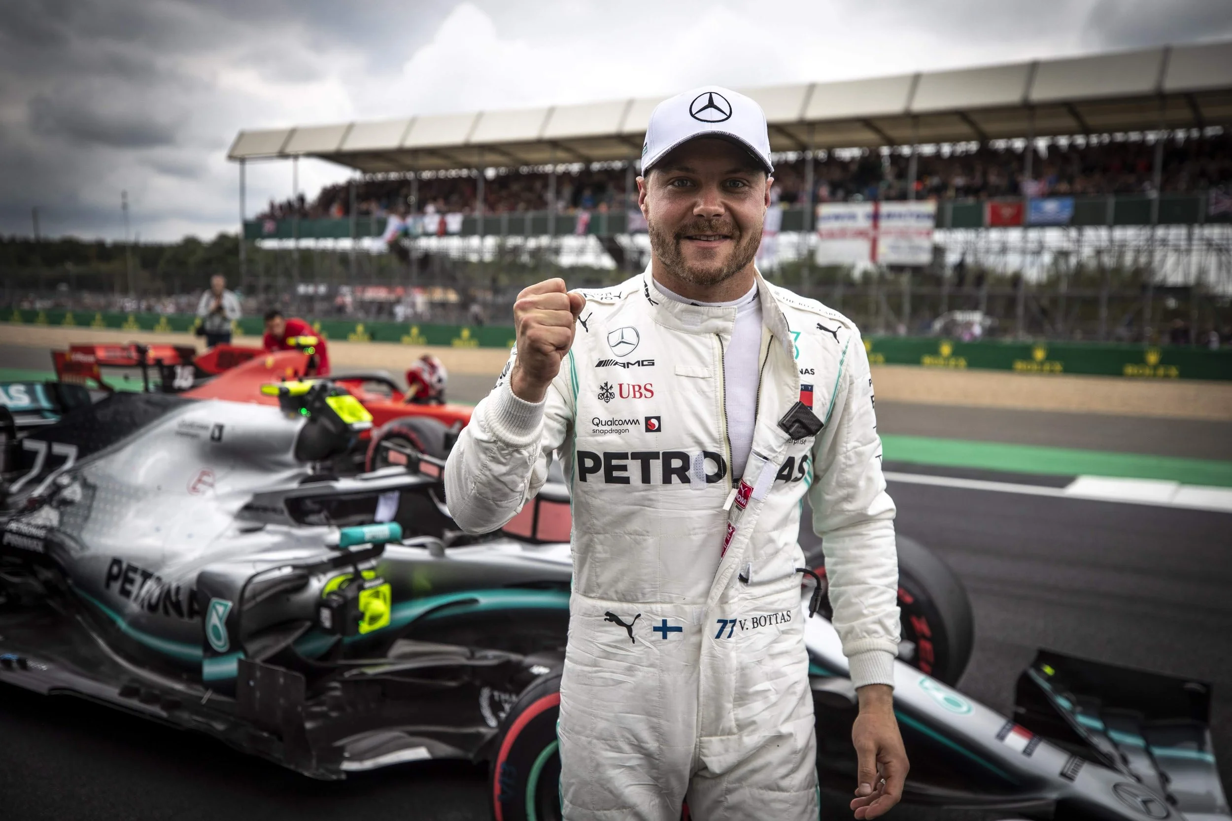 F1 driver Valtteri Bottas celebrating after a race in front of his Mercedes Formula 1 car at the circuit with a grandstand and cloudy sky in the background.