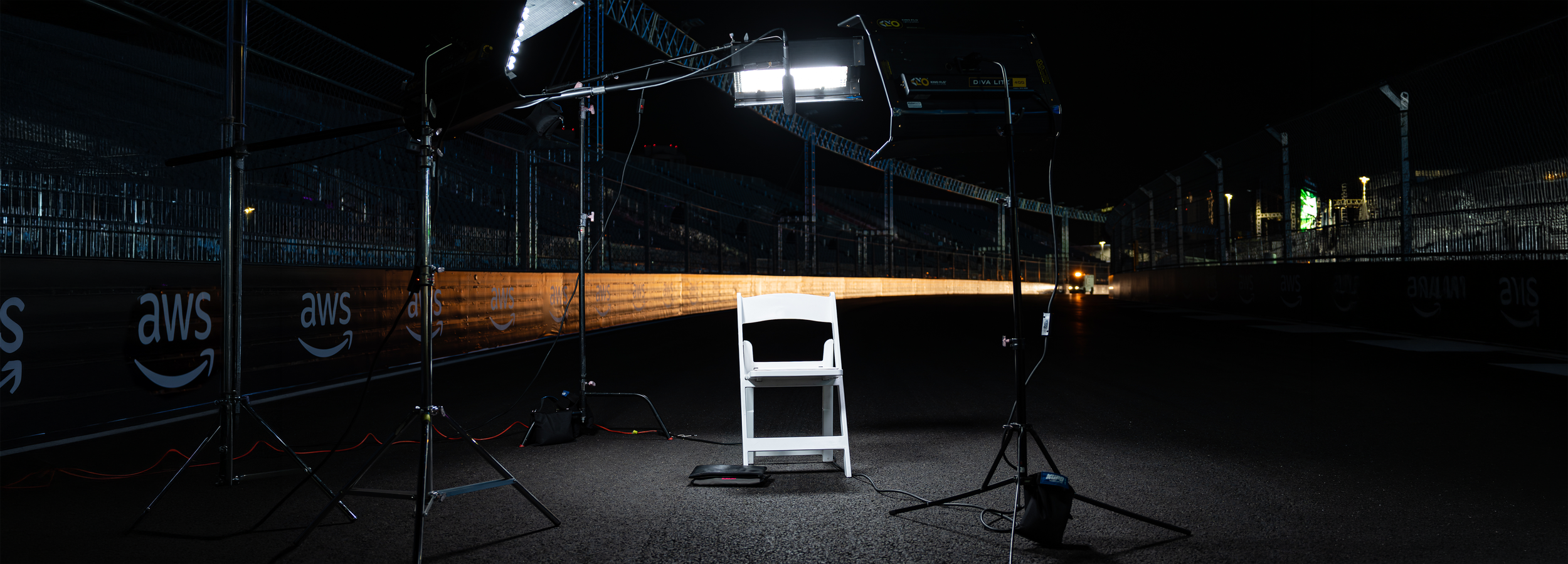 A nighttime scene on a race track with filming equipment, a white chair, and a black bag. The track is illuminated by lights, with branded barriers on the sides.