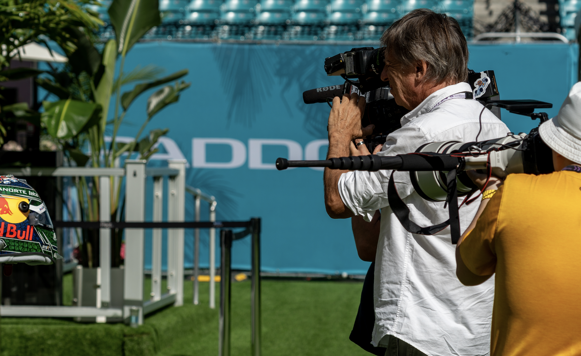 A man with short brown hair, wearing a white shirt, is taking a photograph with a professional camera at a racing event. In the background, there are blue bleachers and a large blue sign, and a person in a yellow shirt is partially visible.
