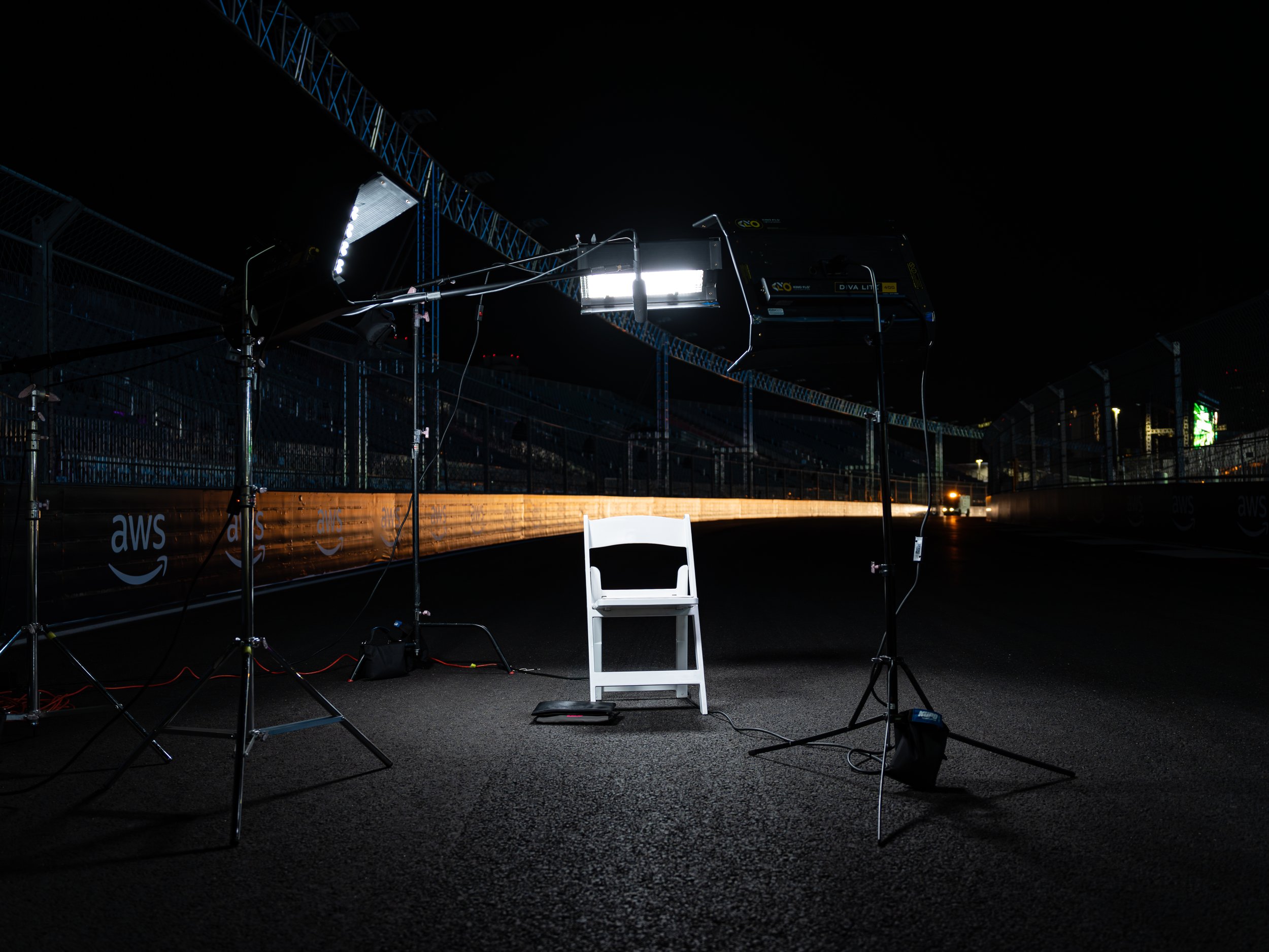 Nighttime scene of a photography or filming setup on a racetrack, with lighting equipment, cables, a white chair, and fencing in the background.