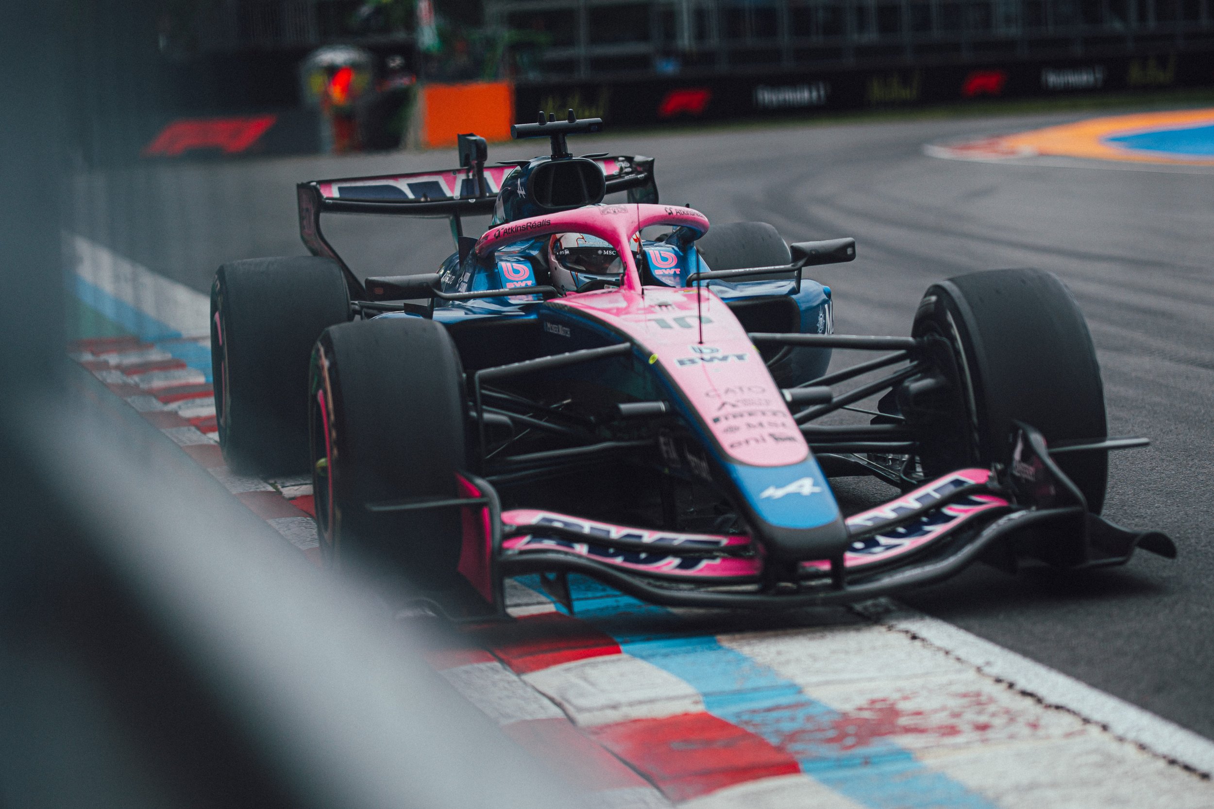A BWT Alpine Formula One car drives through a wet corner on track with spray and blurred barriers in the foreground.