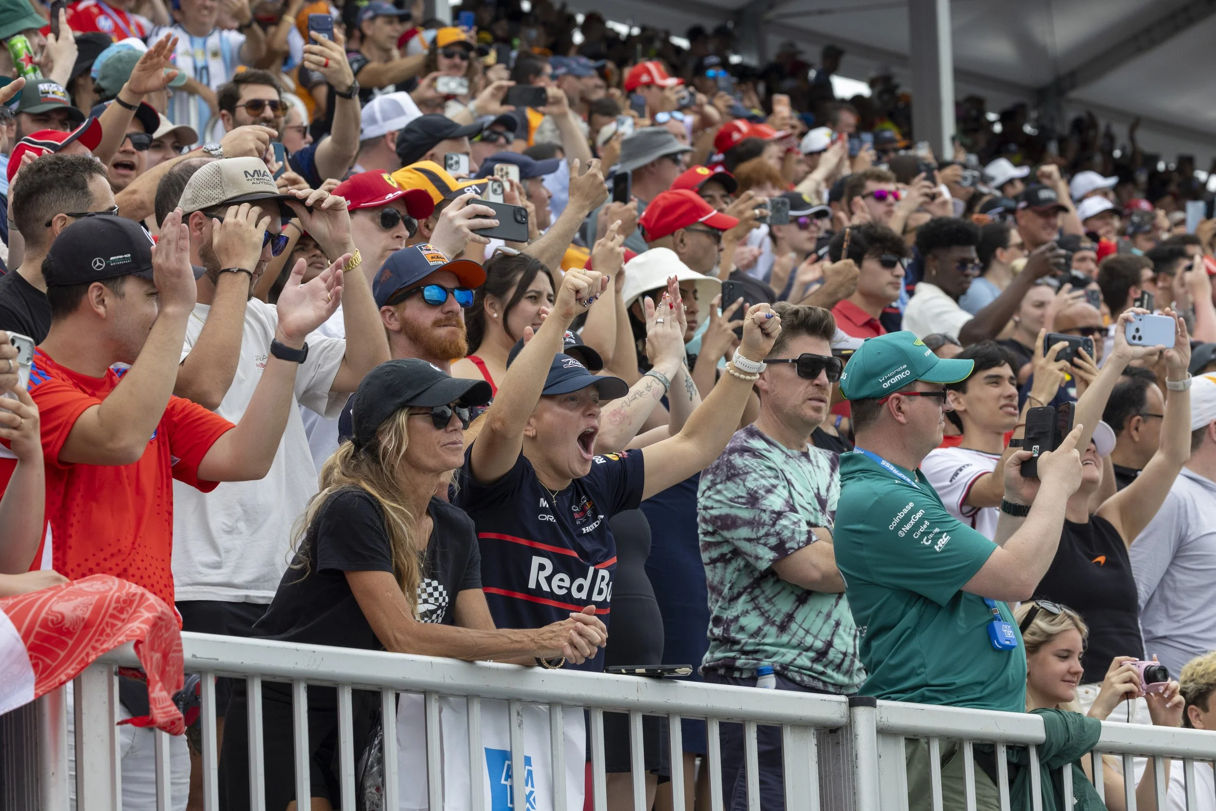 A crowd of spectators at a racing event, with many holding up smartphones to take photos or videos. Some are wearing branded racing team shirts, caps, and sunglasses, and are standing behind a metal barrier.