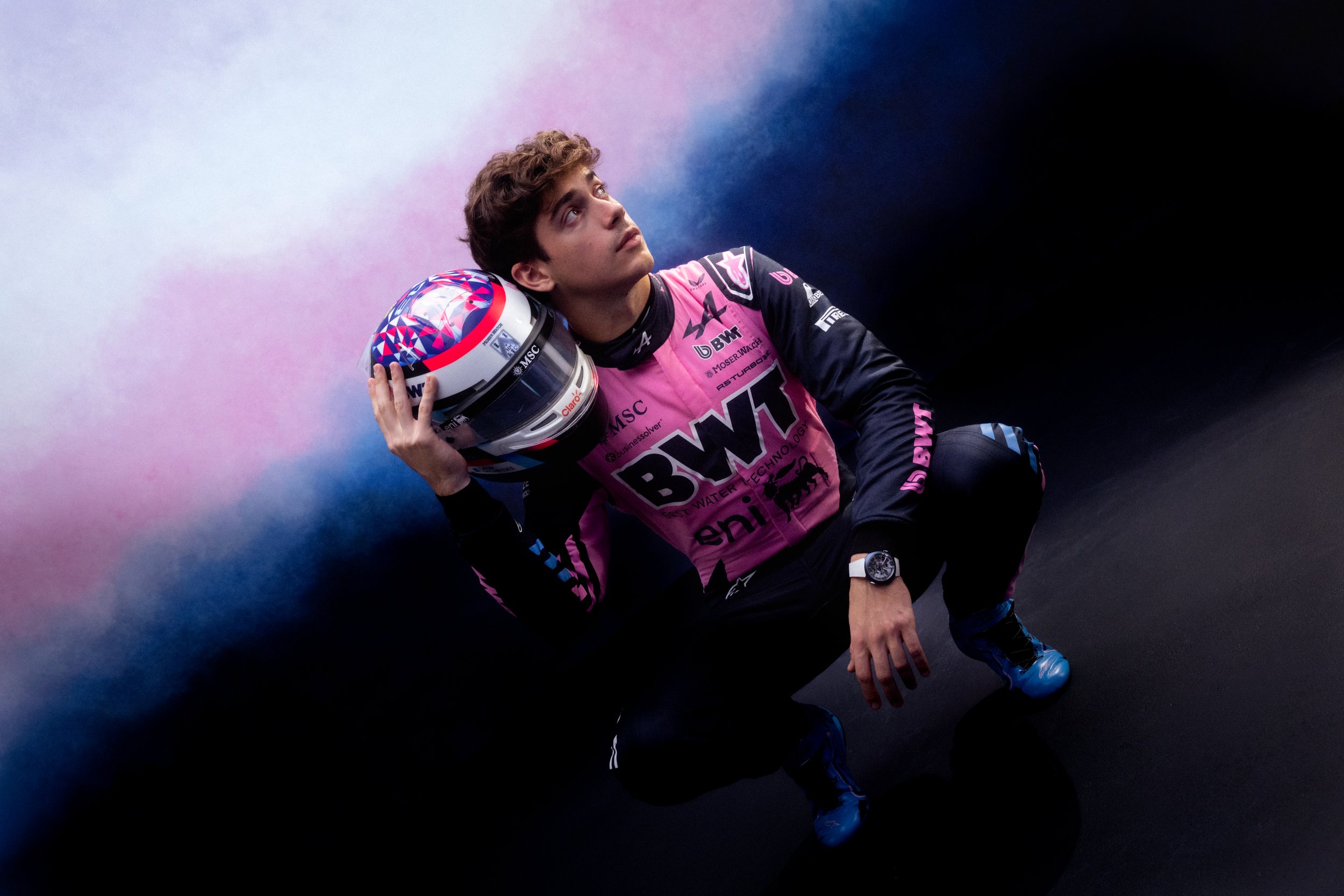 Franco Colapinto poses in BWT Alpine Formula One teamwear against a dramatic pink and blue smoke background.