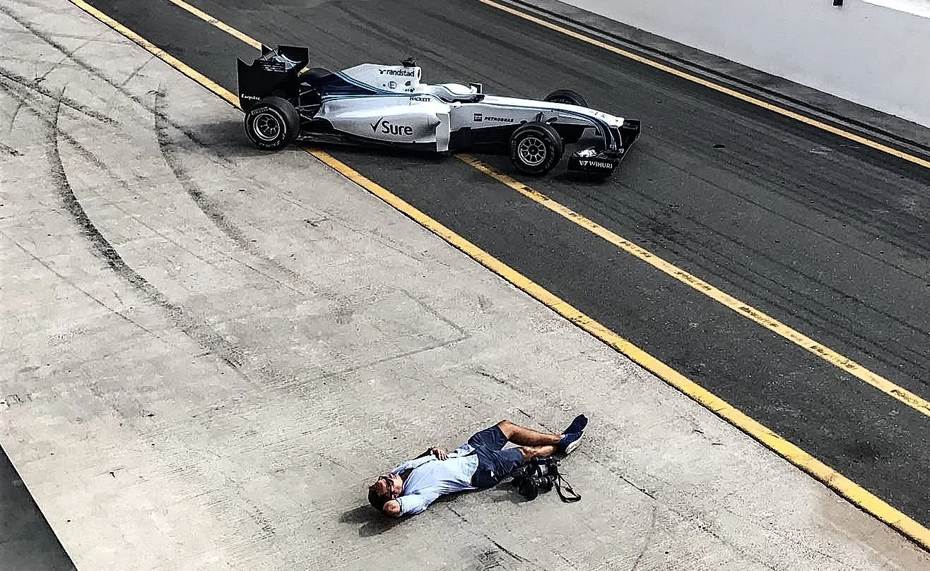 A man in a light blue shirt and shorts lies on the pavement near a race track, with a racing car stopped just a few feet away on the track.