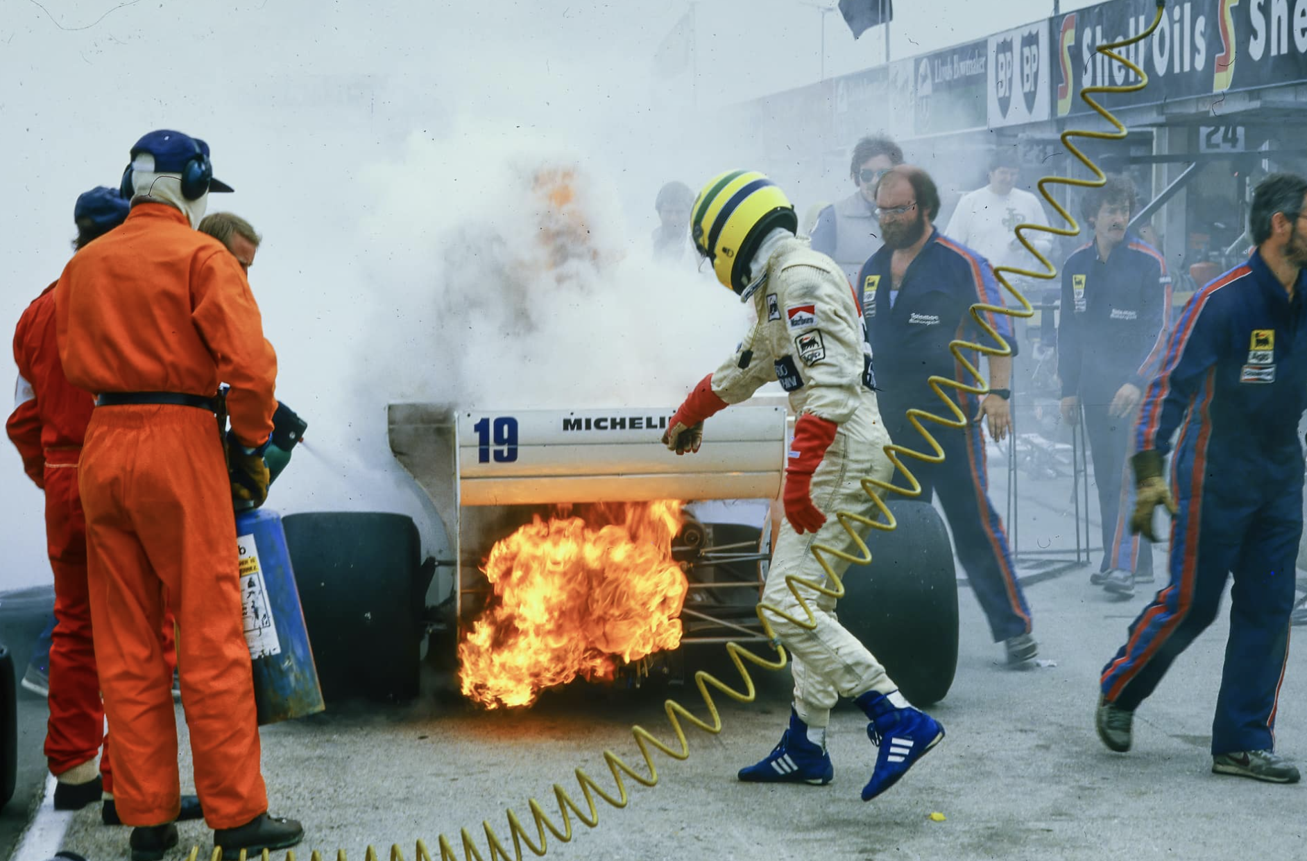 Race car pit crew members handling a car on fire during a race, with one crew member wearing a white racing suit and yellow helmet, and others in blue and orange uniforms nearby.