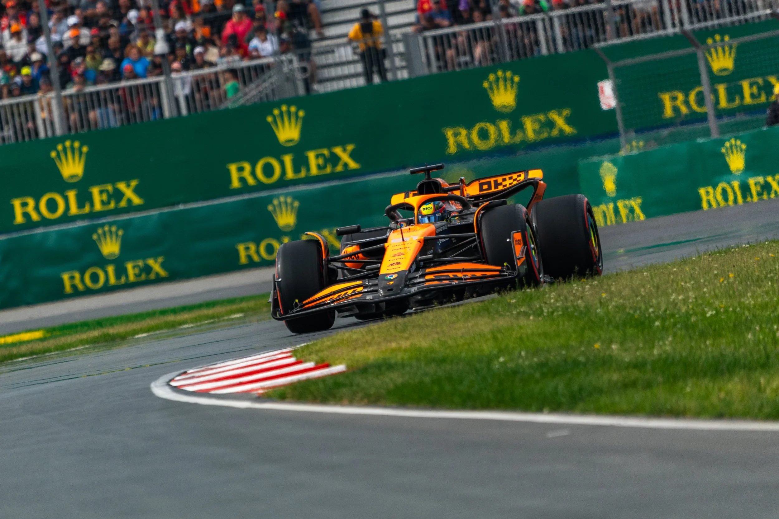 McLaren race car on a turn at a racing circuit with a crowd watching behind green Rolex advertisements.