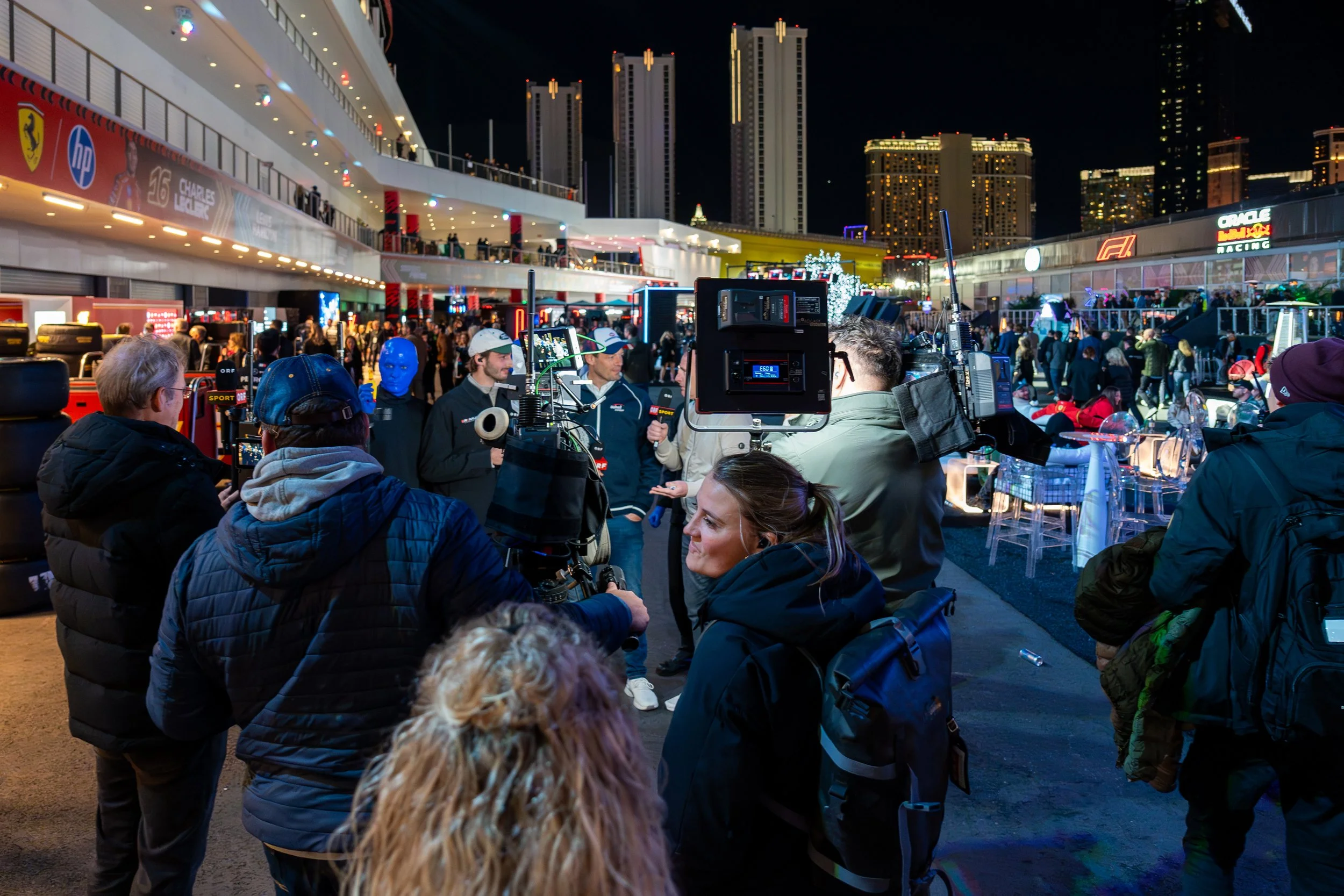 Media crew filming an interview at Formula 1 Las Vegas Grand Prix Paddock event at night with city skyscrapers in the background, and spectators and booths present.