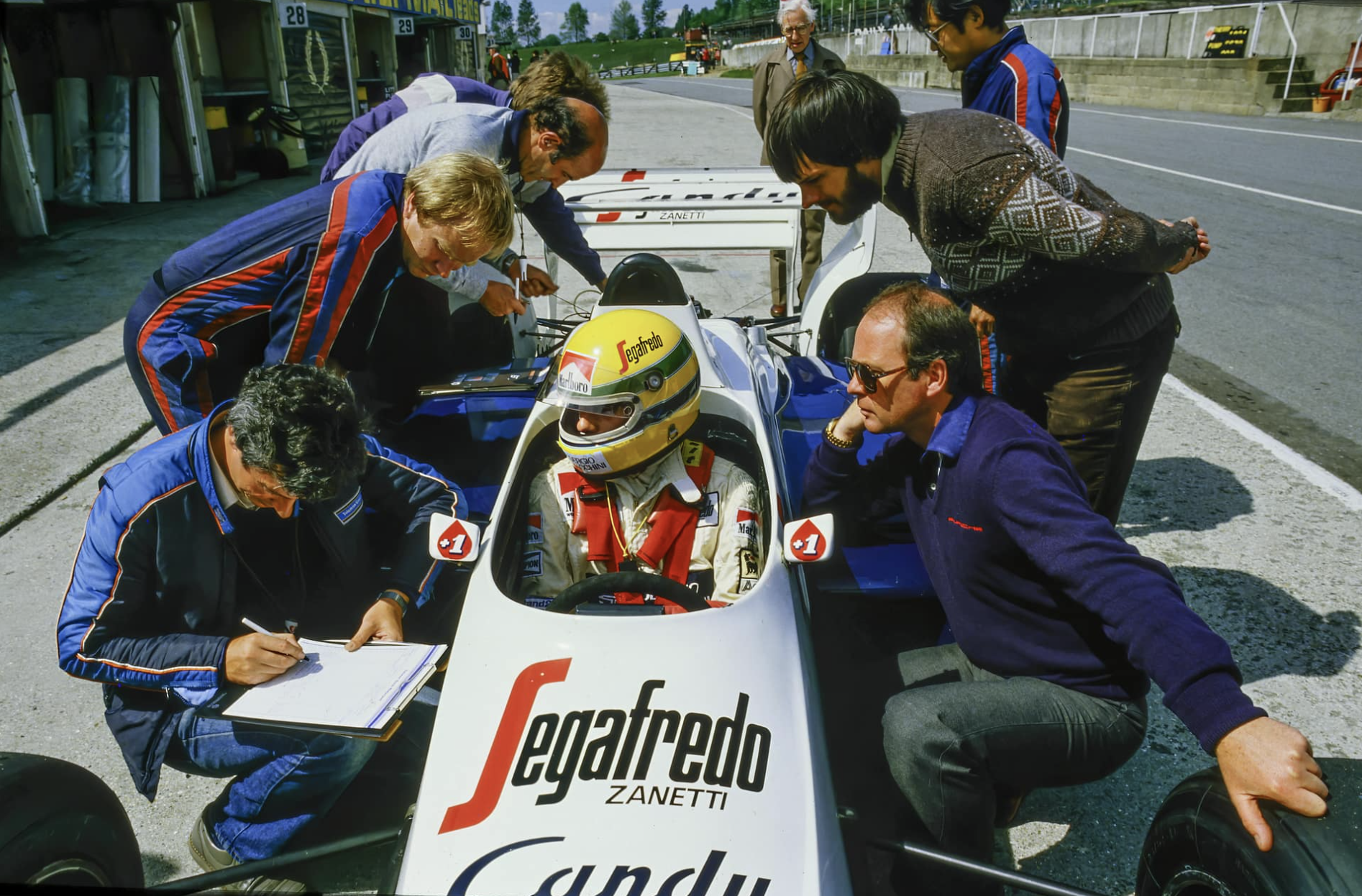 A race car driver wearing a yellow helmet sits inside a Formula 1 car in the pit lane, surrounded by team members and officials. The team members are examining the car, taking notes, and discussing during a race event.
