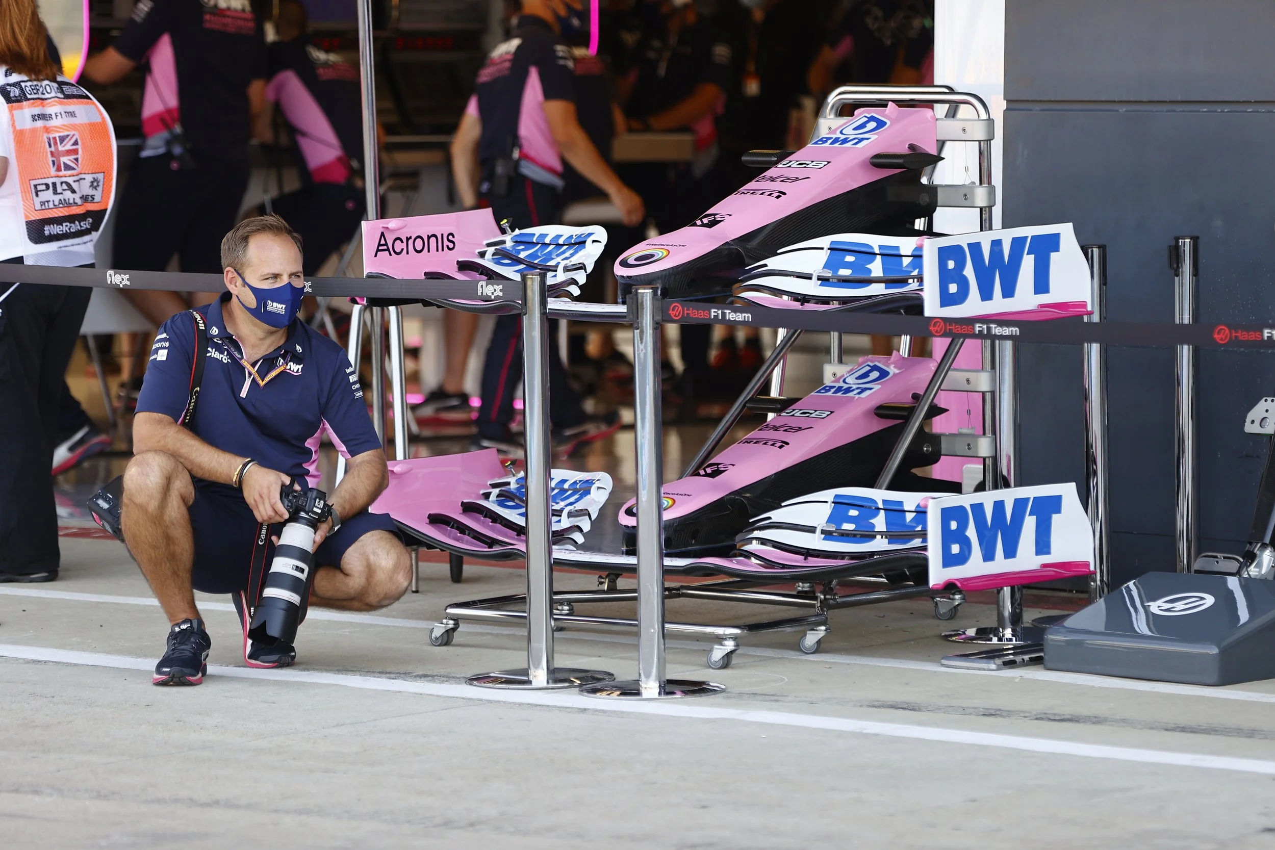 A man crouching in front of a pit stop area for a Formula 1 race car, with two pink and black race cars partially visible behind a barrier. The man is wearing a blue face mask, a blue team shirt, black shorts, and holding a camera with a large telephoto lens. Other team members in pink and black shirts are visible in the background.