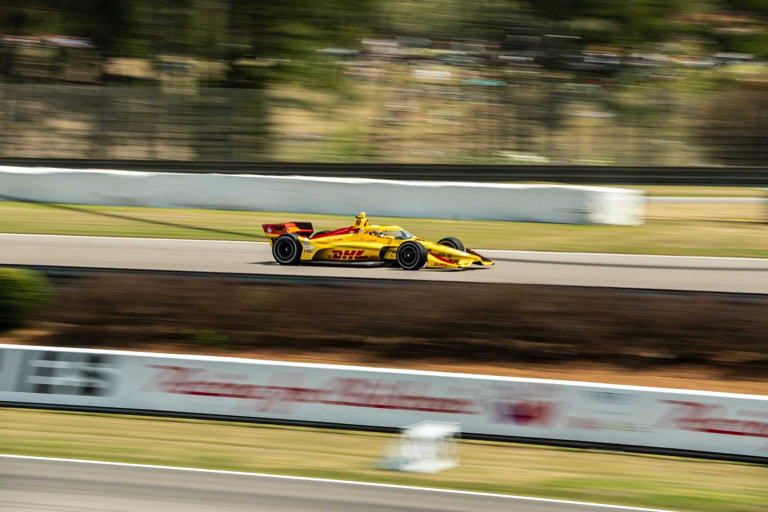 A yellow and red racing car speeding on a track with blurred background and fencing, indicating high-speed motion.
