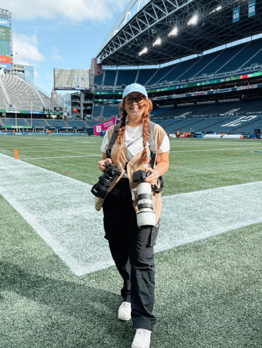 Hannah Palpant standing on a football field at a stadium, holding a camera with a large telephoto lens, wearing a white cap, beige vest, white t-shirt, black cargo pants, and white sneakers.