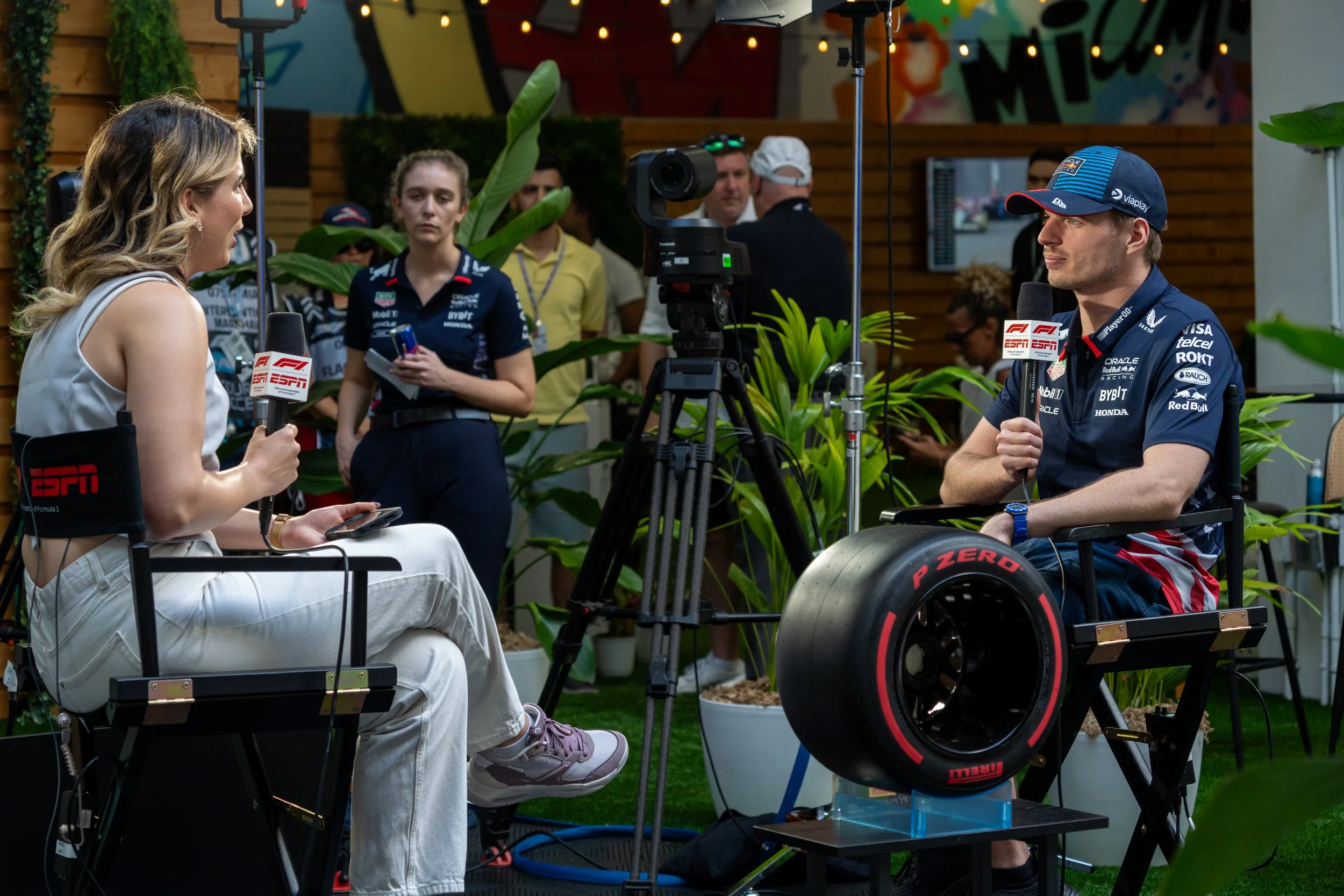 An interview with a woman and a man in a wheelchair, both holding microphones with ESPN logos, is taking place outdoors. The woman is sitting on the left, wearing a sleeveless white top and white pants, holding a smartphone. The man is sitting on the right, wearing a racing team uniform and cap, with a racing wheel attached to his wheelchair, and looking at the woman. Several people are observing in the background, and the setting is decorated with green plants and colorful graffiti on the wall.