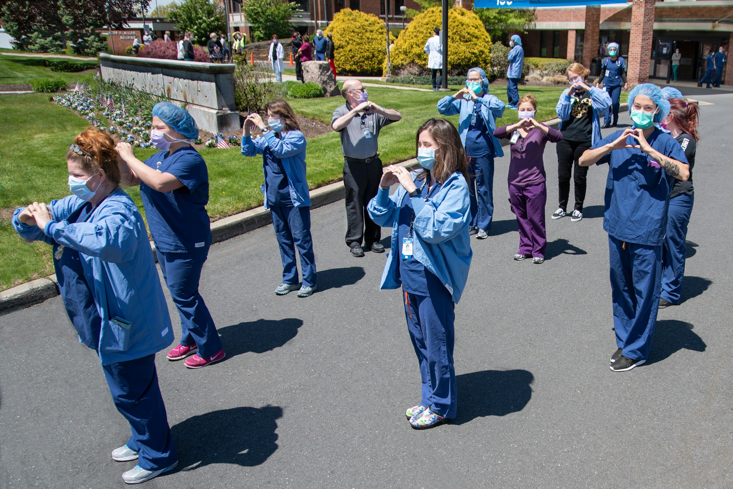 Healthcare workers standing outdoors, wearing scrubs and face masks, practicing hand gestures, with a building and greenery in the background.