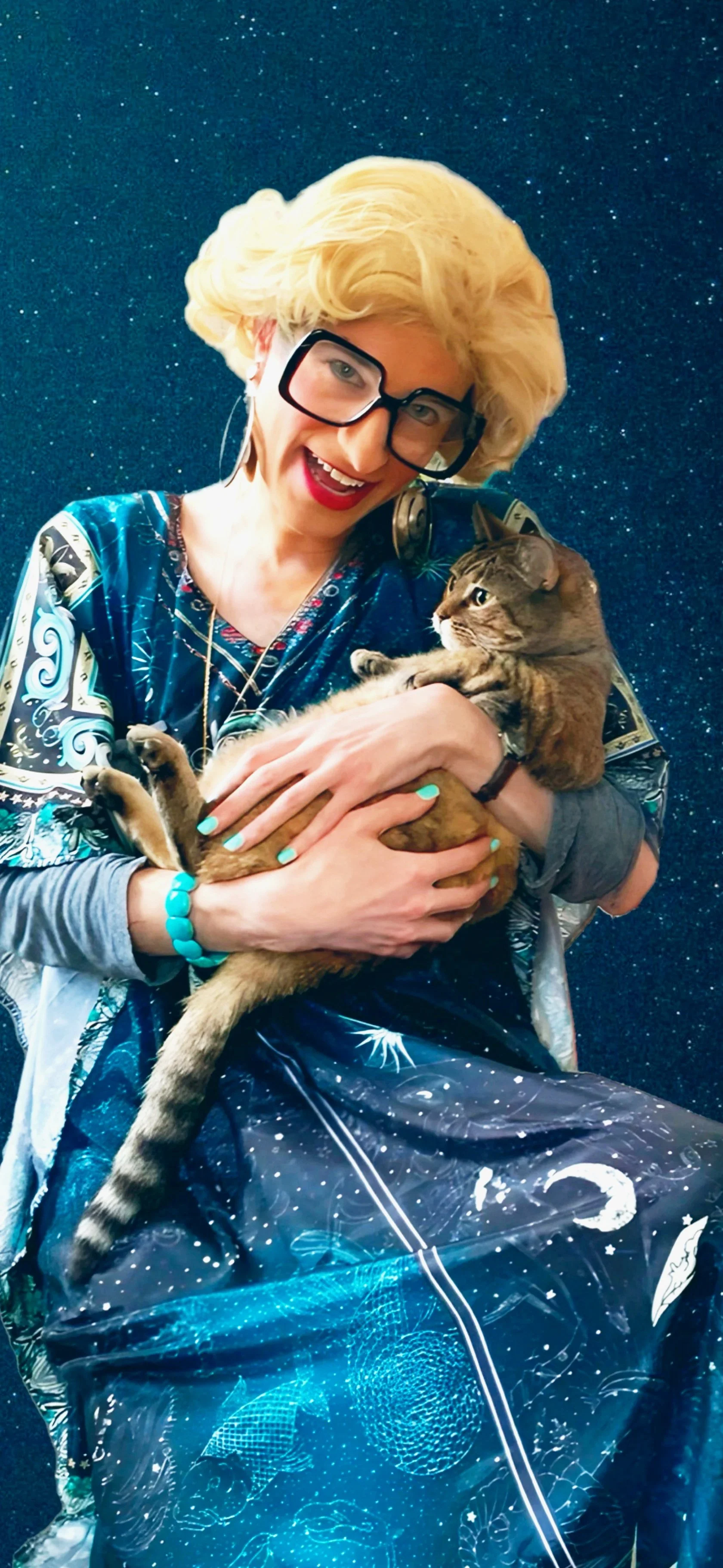 A woman with blonde hair, glasses, and red lipstick smiling while holding a tabby cat, in front of a starry background.