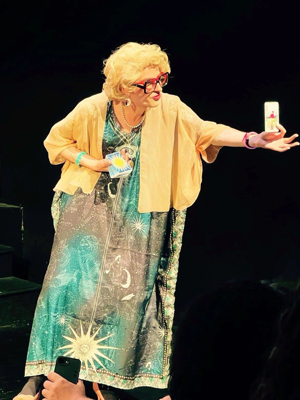 Person with blonde curly hair, red glasses, and colorful attire taking a selfie on stage.