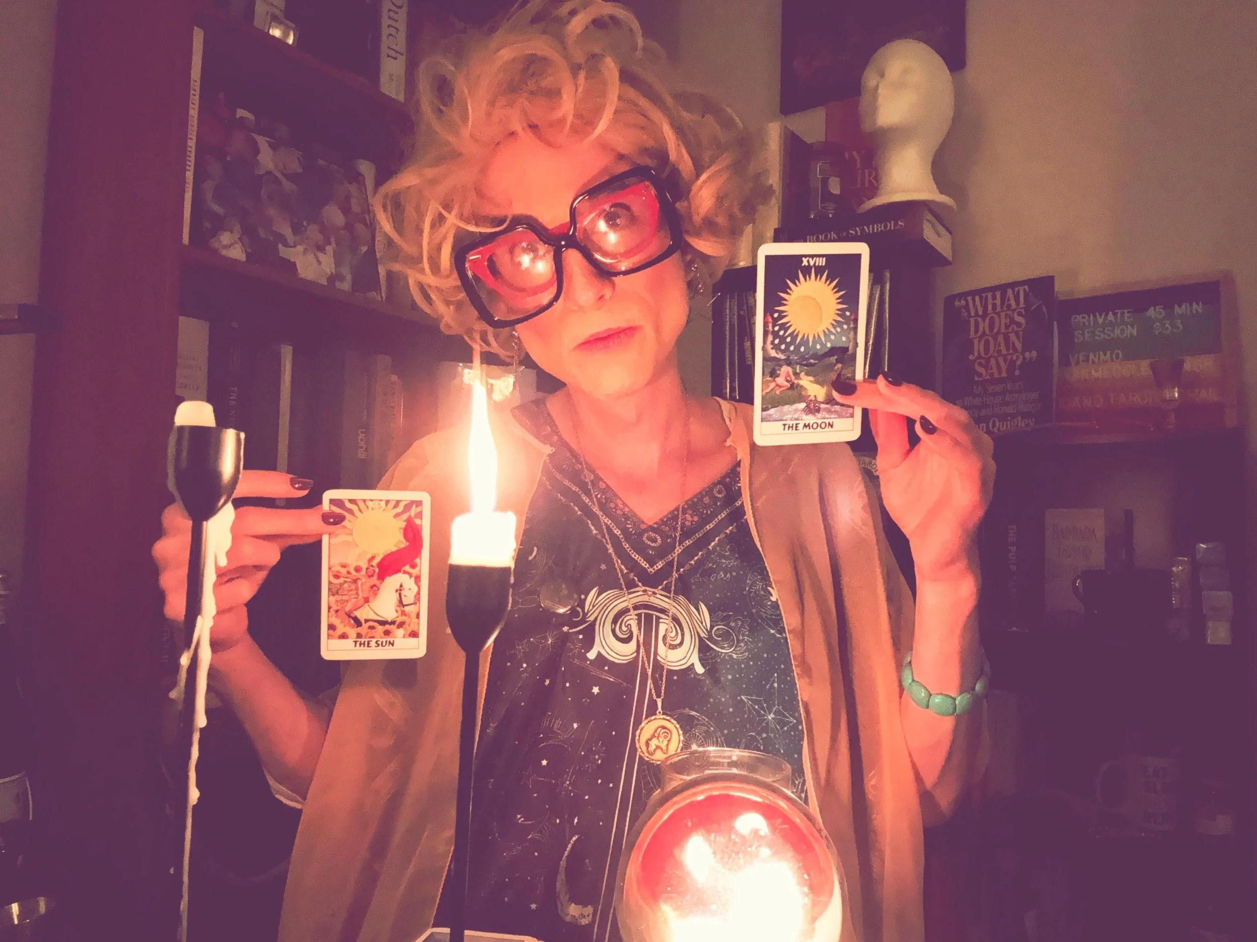 Woman with curly blonde hair, wearing large pink glasses, holding two tarot cards, the Sun and the Moon, in a dimly lit room with candles.