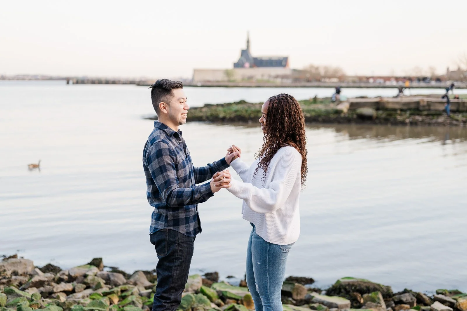 A couple holding hands and smiling at each other near a body of water with rocks along the shore and a building in the background. Couple photography. Portrait photography.