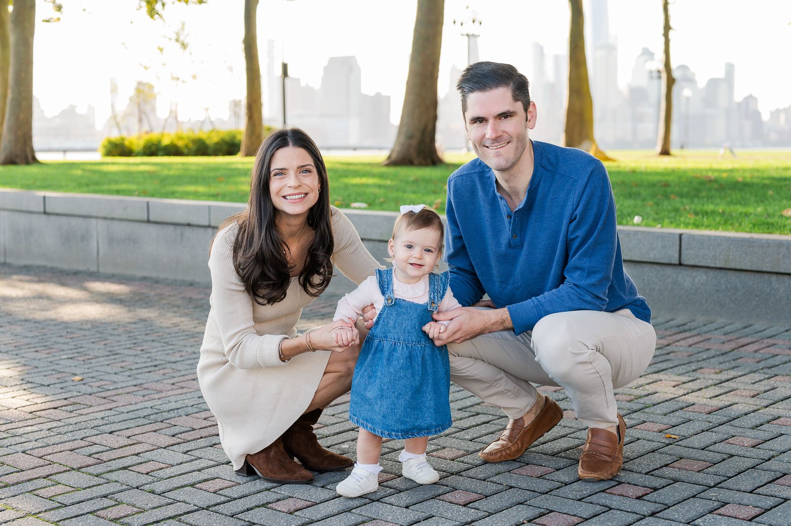 A happy family of three, a woman, man, and a girl, smiling in a park with a city skyline in the background. Family photography. Portrait photography.