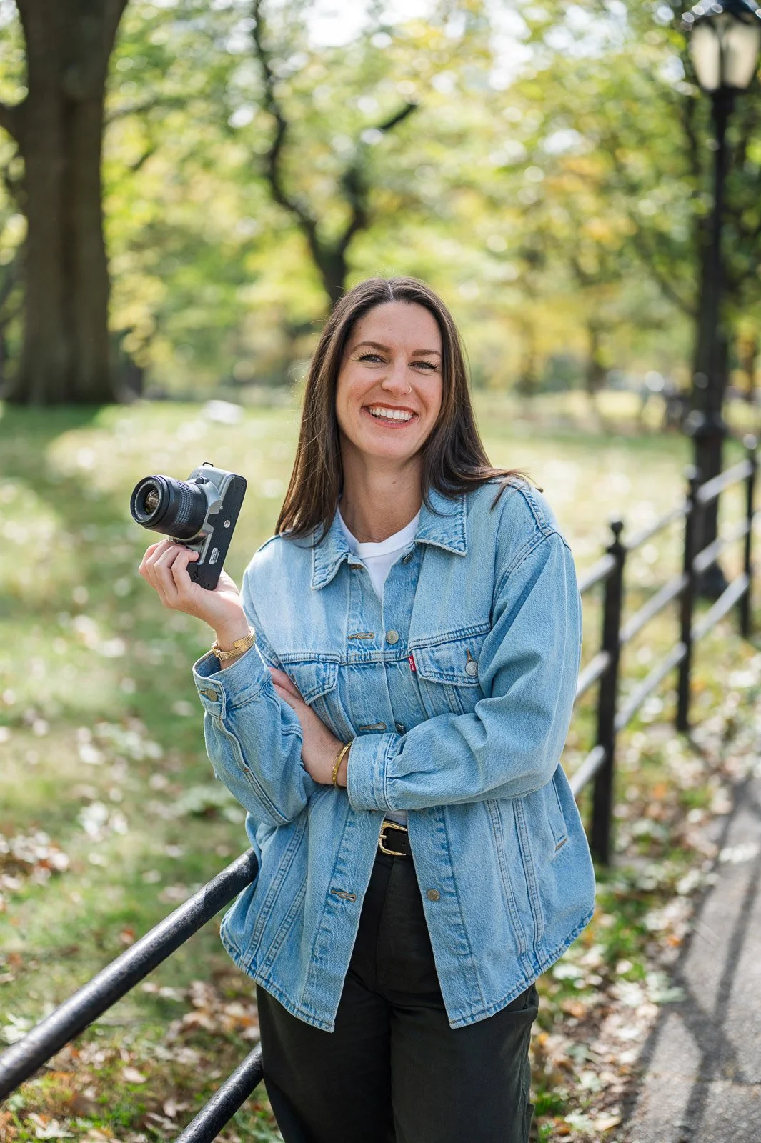 A woman with long dark hair, wearing a denim jacket, smiling while holding a camera in a park with green trees and fall foliage.
