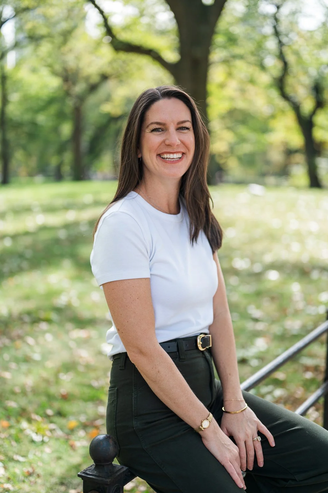 A woman with long brown hair smiling, sitting outdoors on a park bench with trees and greenery in the background.