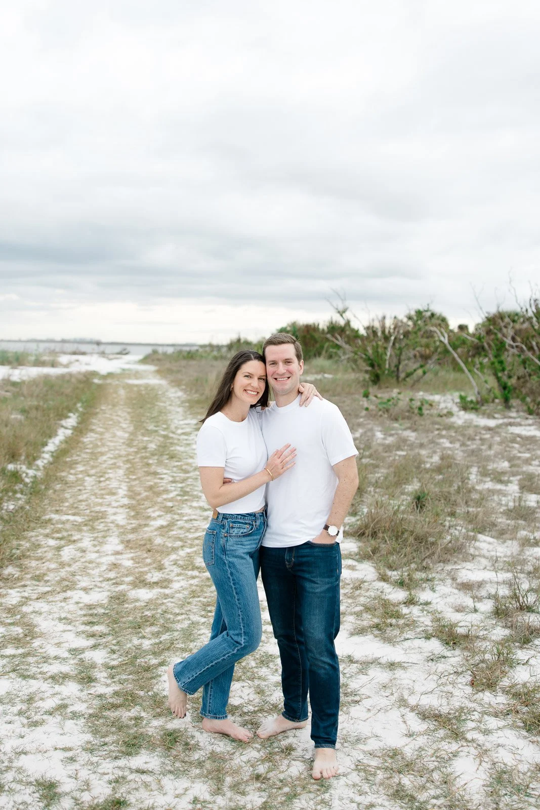 A smiling couple stands barefoot on a sandy path in a beachy area, hugging each other, with overcast skies and sparse greenery in the background.