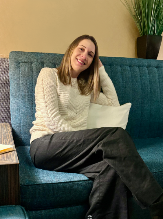 A woman with shoulder-length hair sitting on a teal sofa, smiling, in a cozy indoor setting with a beige wall, a white pillow, a wooden side table, and a tall green potted plant.