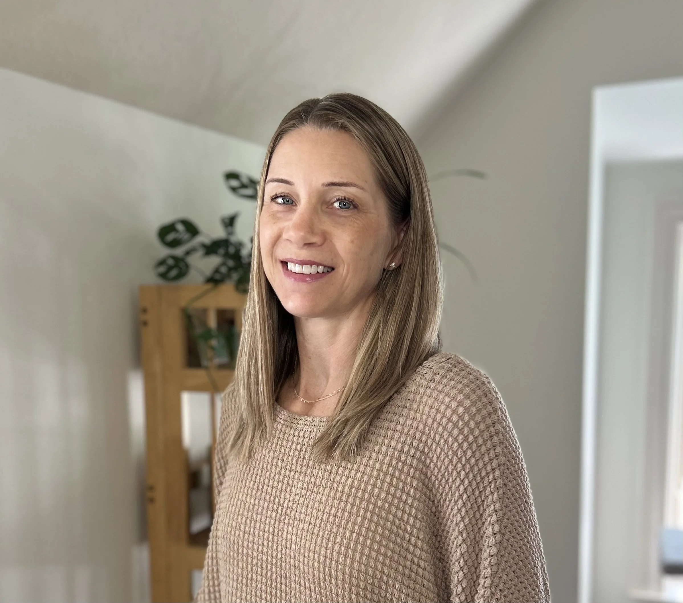 A woman with brown hair and blue eyes smiling in a beige sweater, standing inside a house with light-colored walls and a plant in the background.
